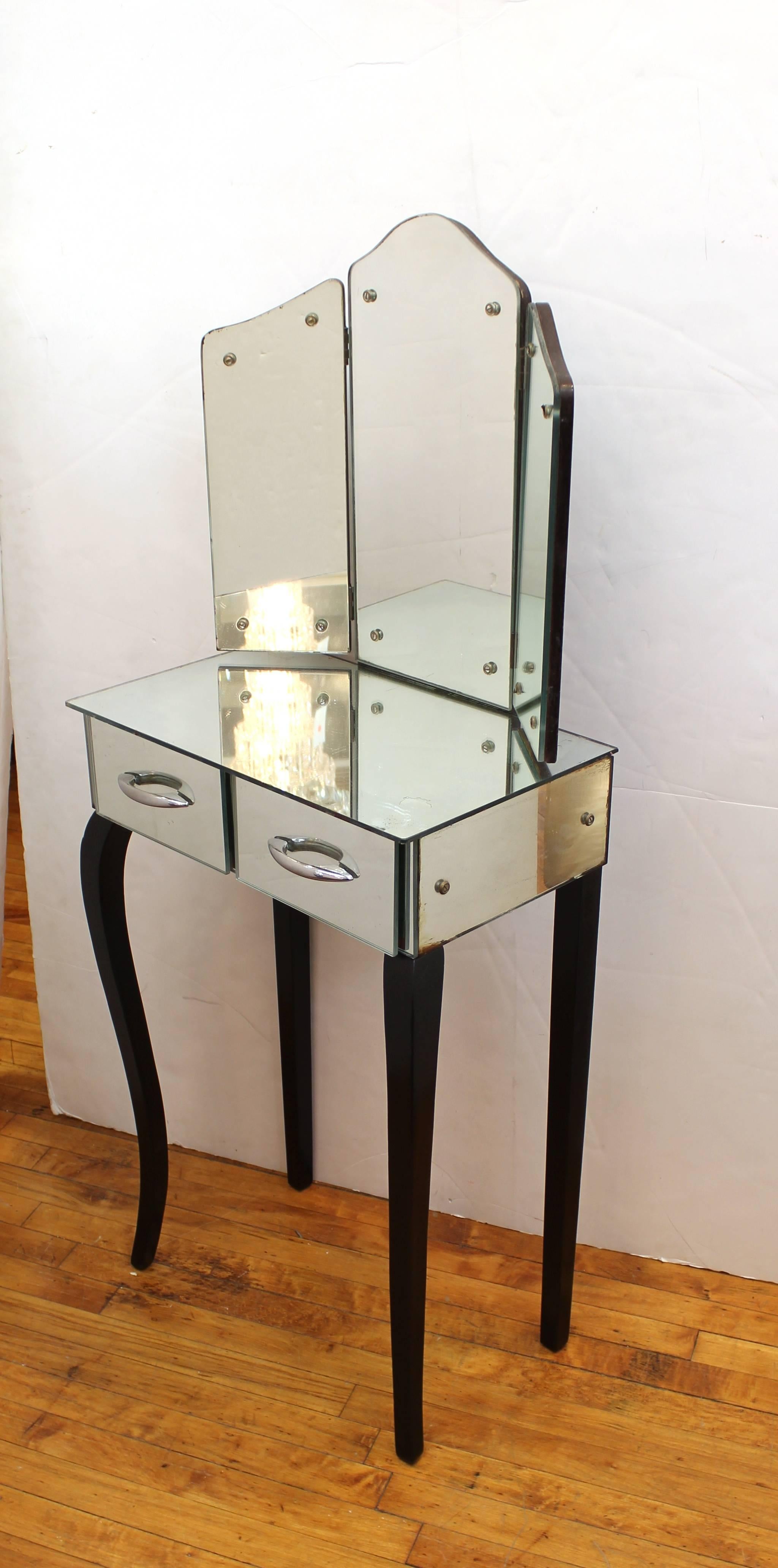 Art Deco vanity table with trifold mirror produced in France during the 1930s. The table features a mirrored surface including two drawers. A trifold mirror with arched center panel is also included. The piece sits on gently curved black legs. Wear