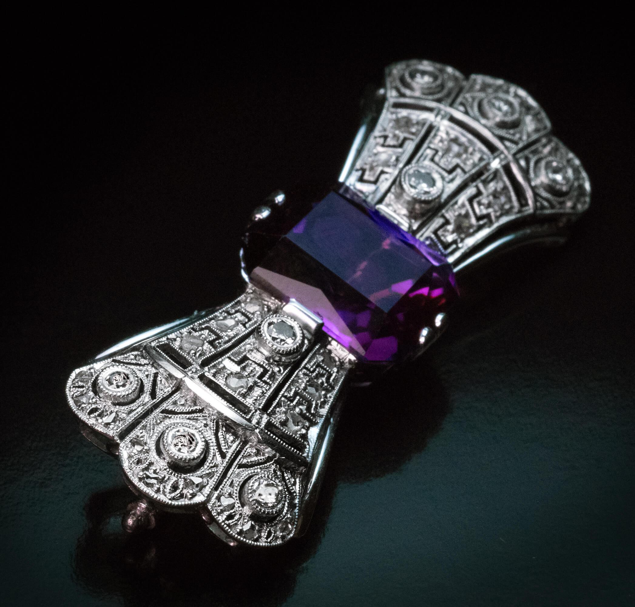 Central Europe, circa 1925  The bow shaped brooch is handcrafted in 14K white gold and centered with an octagon mixed cut amethyst (13.68 x 11.16 x 7.19 mm, approximately 7.27 carats) of a deep purple color with some flashes of pink. The openwork