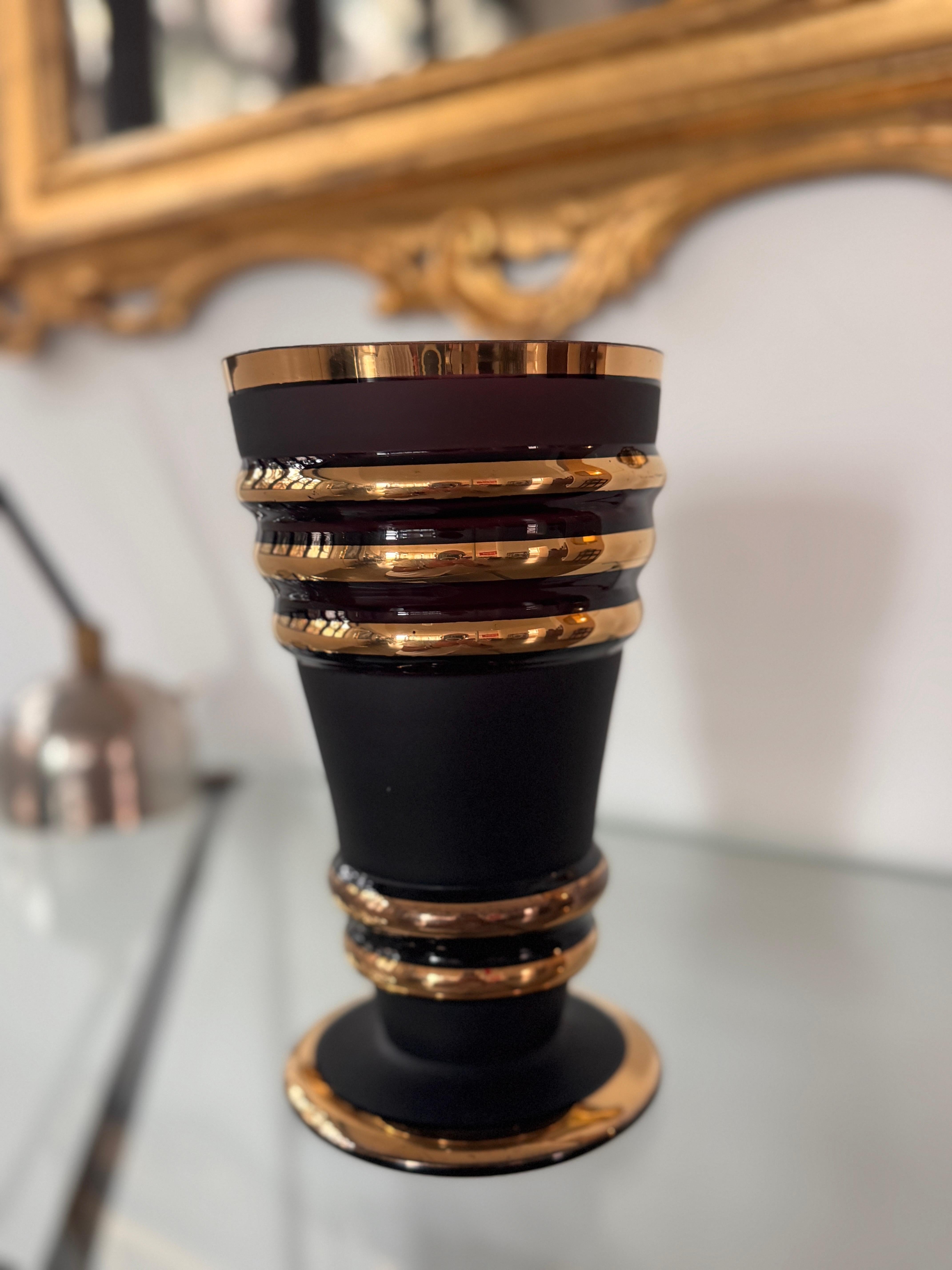 Elegante Vase aus mundgeblasenem Glas in tiefem Pflaumenton (dunkler Amethyst), verziert mit feinen Goldbändern und -ringen.
Der Kontrast zwischen der matten Oberfläche und der leuchtenden Vergoldung verleiht dem Stück eine markante