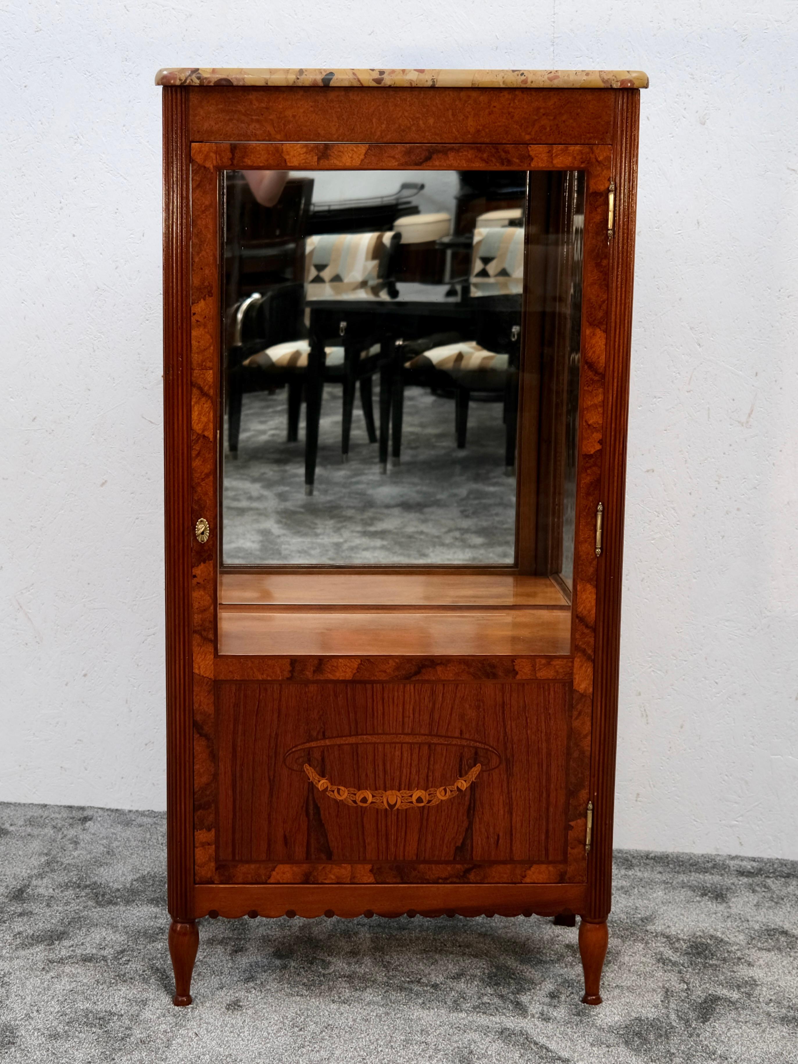 A refined and compact Art Deco display cabinet from around 1925, crafted in richly grained amboyna wood and crowned with a polished marble top in warm, earthy tones. The front and sides are glazed, providing a clear stage for treasured objects, with