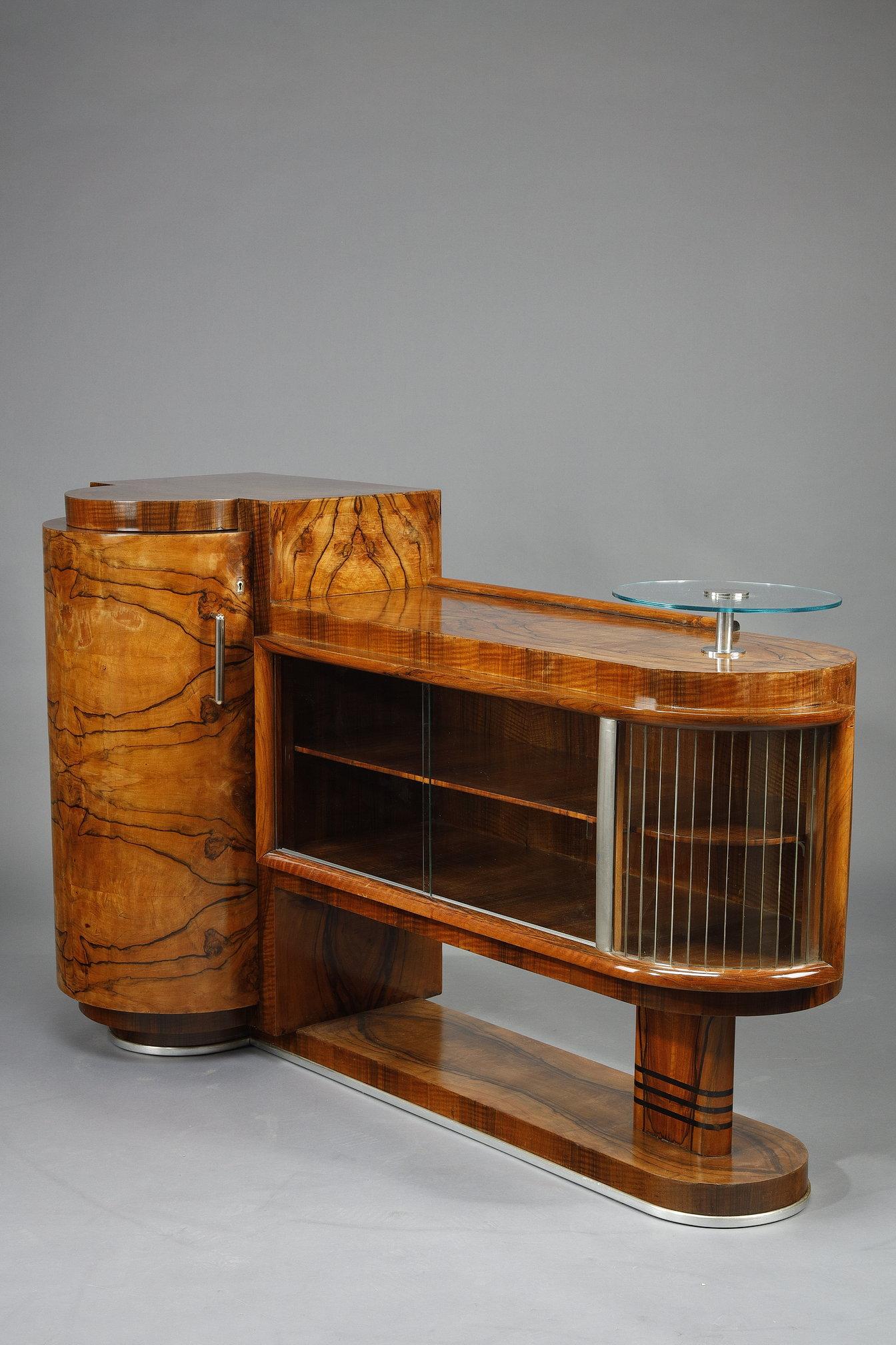 Credenza d'epoca Art Déco in noce e impiallacciatura di noce che si apre su un'anta cilindrica e porte scorrevoli in vetro. Base e maniglie cromate. Un piano circolare in vetro. La sua forma architettonica evoca le forme e le linee moderniste