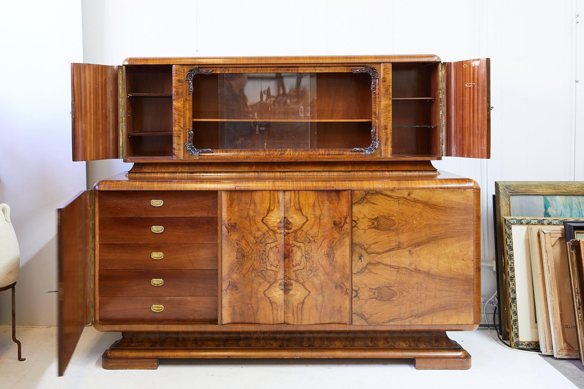 Early 20th century German Art Deco period buffet with upper bar cabinet. An upper bar cabinet features a display behind sliding glass doors and is framed by decorative carved corner moldings. This is flanked by bookmatched burl walnut veneered doors