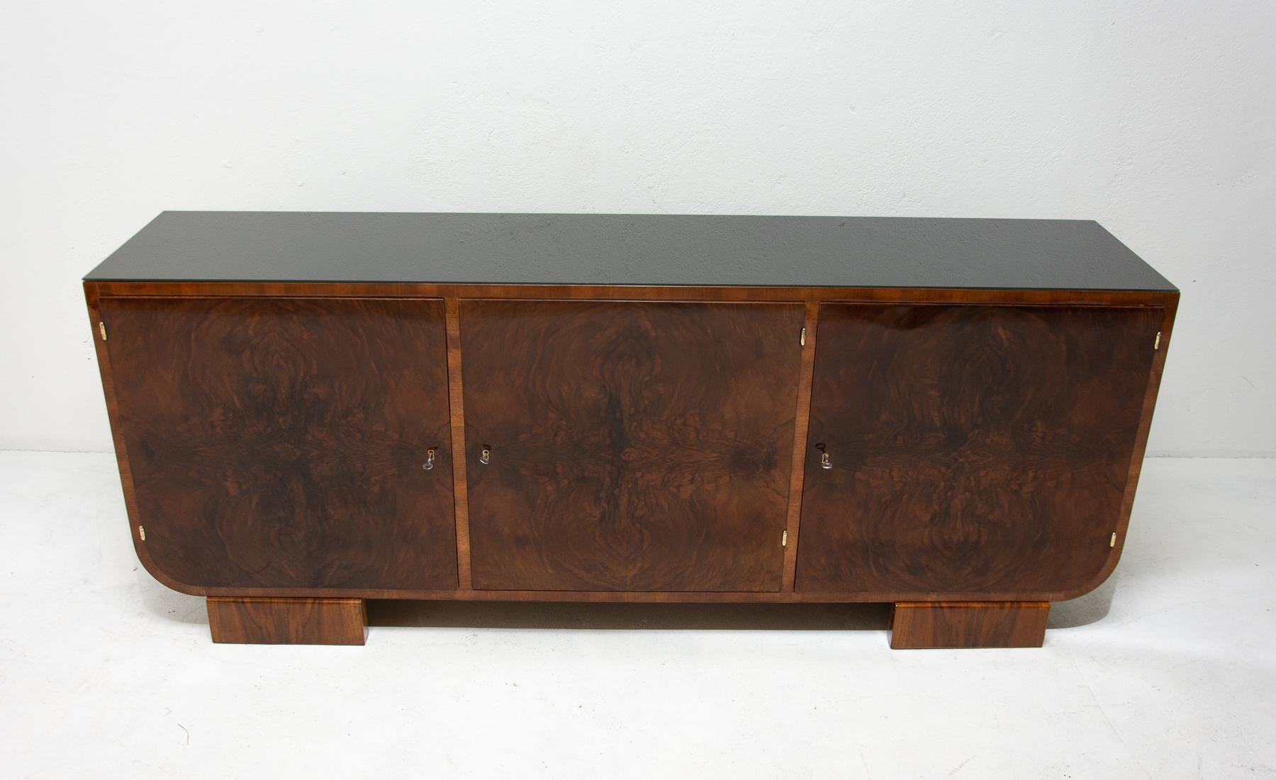 This sideboard was made in Bohemia in the 1930s. It´s an example of Central European functionalism and is made of solid wood and veneered in walnut. It features a storage space divided to three parts and the top is covered with a black glass. The