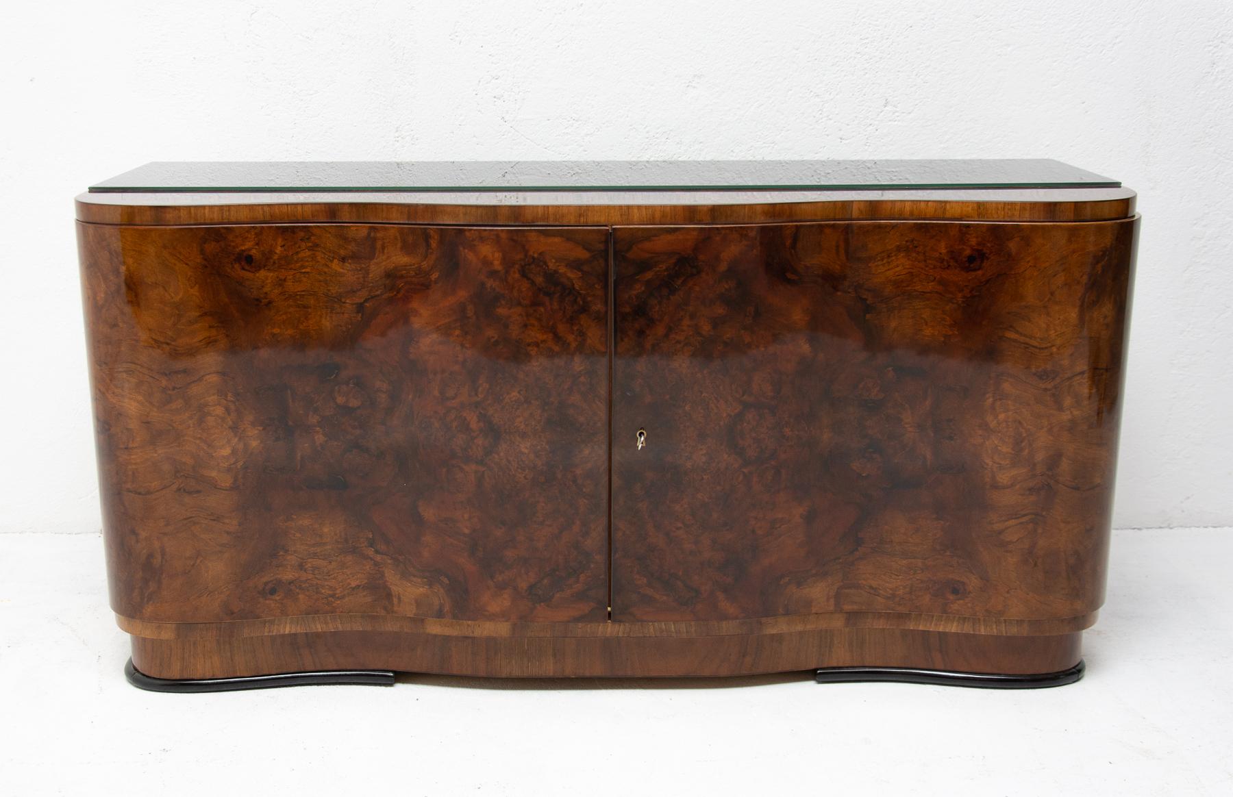 This sideboard was made in Bohemia in the 1930s. It´s an example of Central European Art Deco and is made of solid wood and veneered in walnut. It features a large storage space and the top is covered with a black glass. The cabinet can be also used