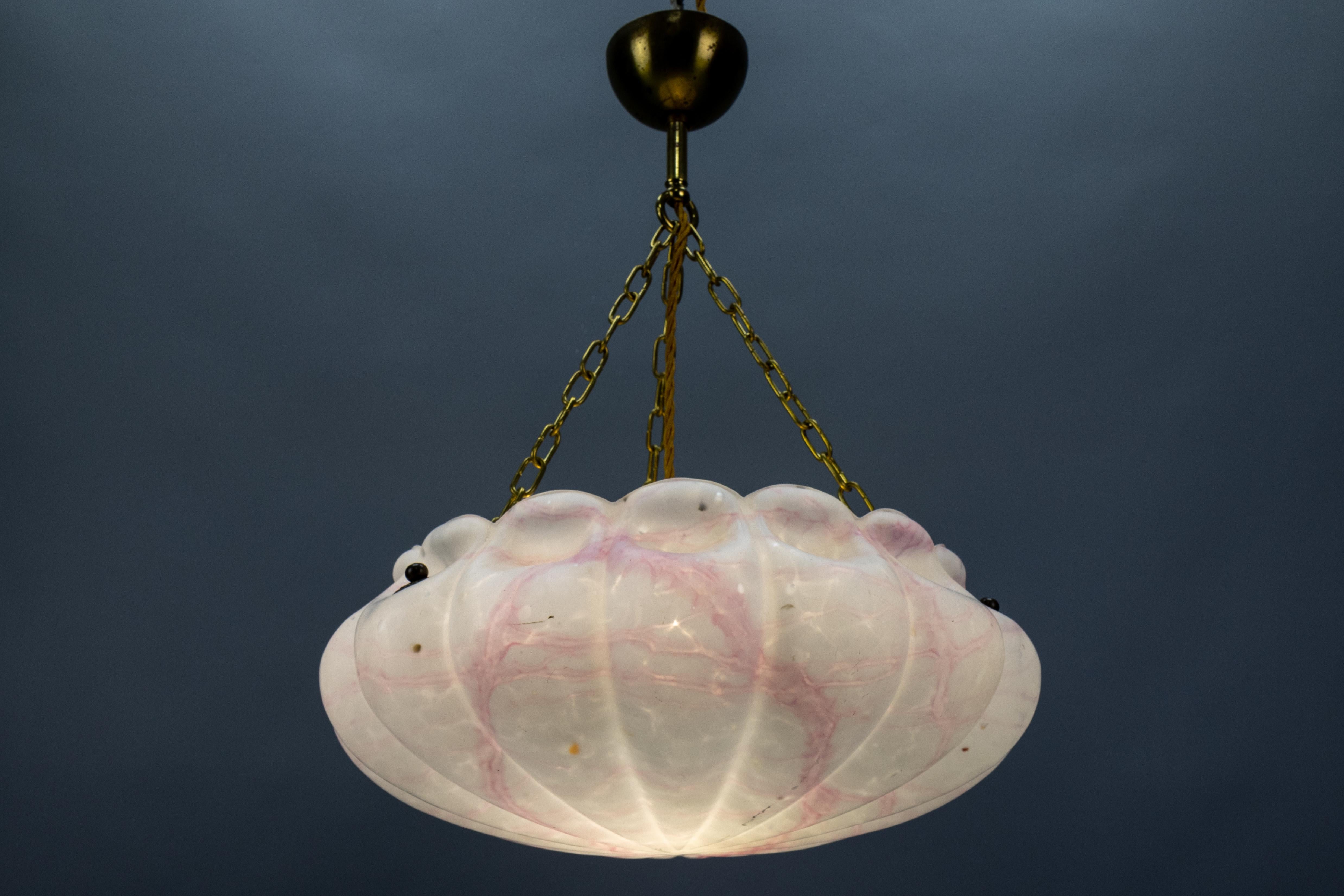 An Art Deco style marbled white and light pink glass and brass pendant light from the 1930s, Germany.
This adorable ceiling light fixture features a beautifully shaped marbled glass lampshade in white and with light pink veins. The glass bowl is