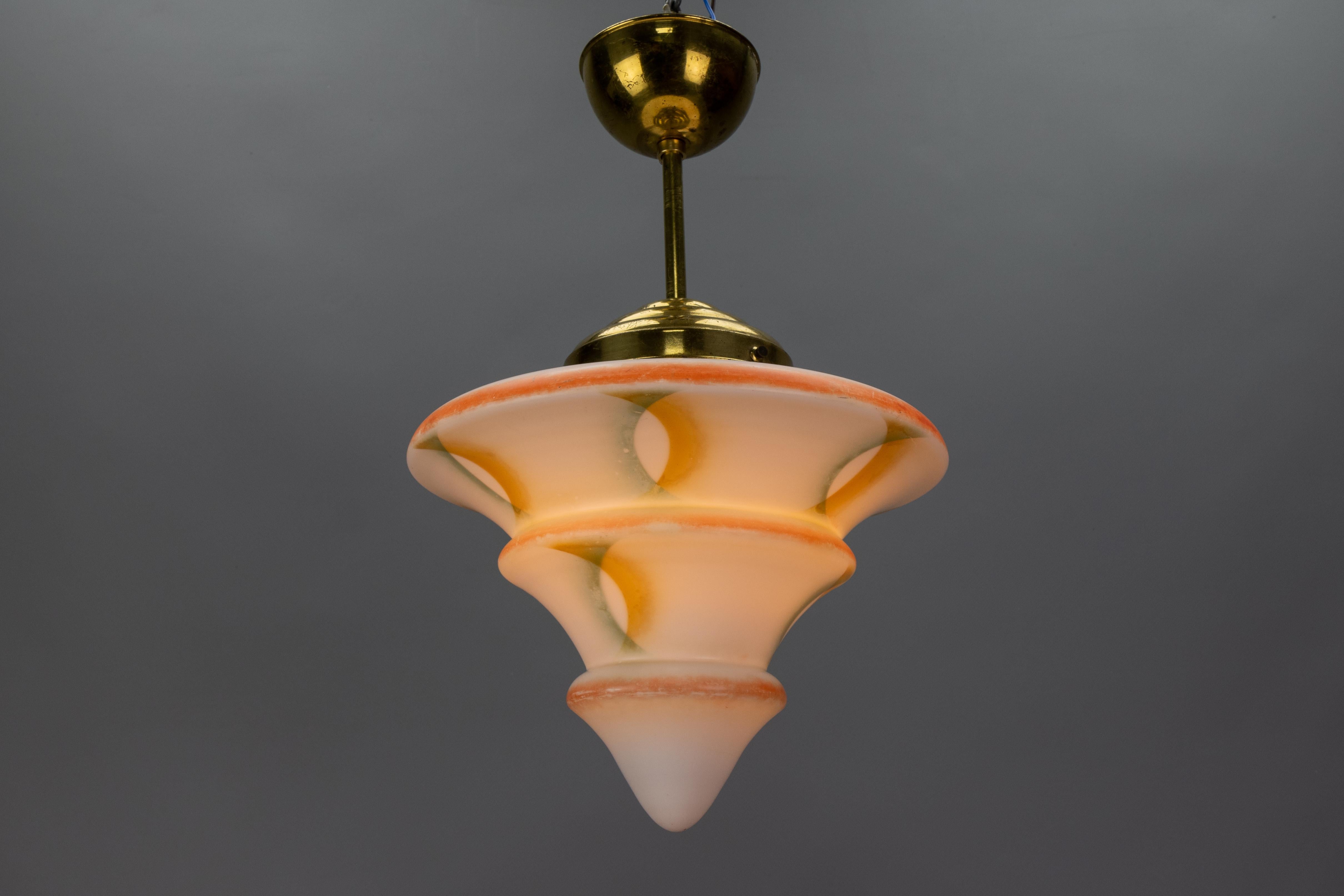 This stunning Art Deco pendant light, crafted in Germany in the 1930s, features an elegant, beautifully shaped white glass shade adorned with decorative accents in green, orange, and salmon colors. The shade is suspended from a brass canopy and