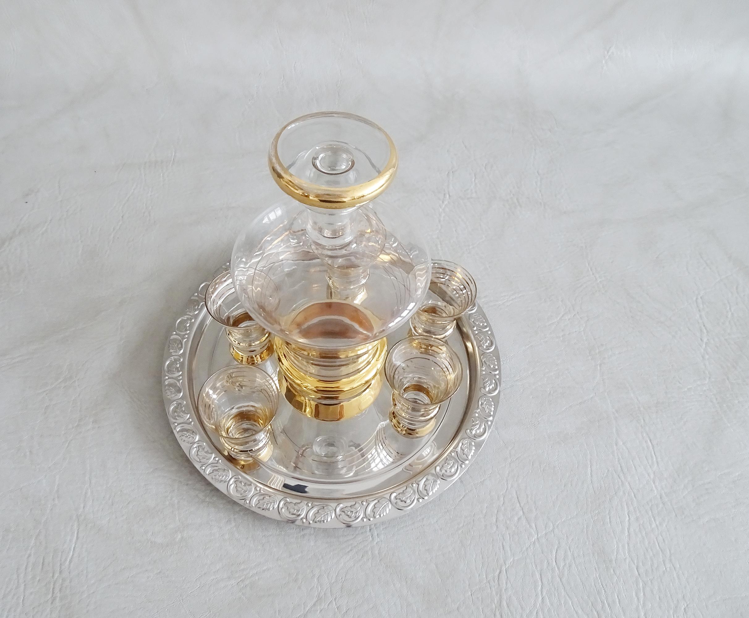 Elegante juego de bar compuesto por una jarra de licor con cinco vasos y una bandeja. Un elegante juego de licor que convence por su color y diseño. Jarra de cristal con rayas doradas y copas de licor a juego. La bandeja redonda de metal para servir