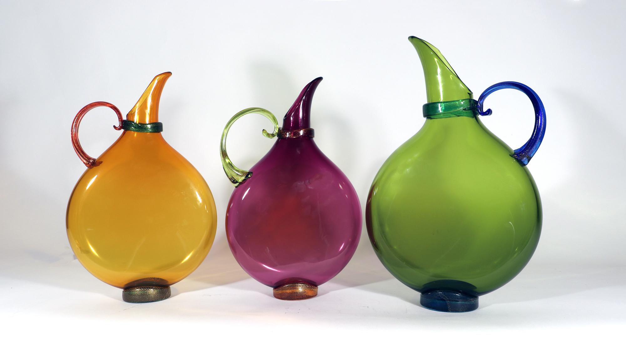 Art glass hand blown flat transparent glass pitchers,
Set of Three.
Nine Iron Studios, Pa.

The grouping of three Nine Iron Studios hand blown flat transparent art glass pitchers in amethyst, topaz, and olive. Each are raised on a gold flecked