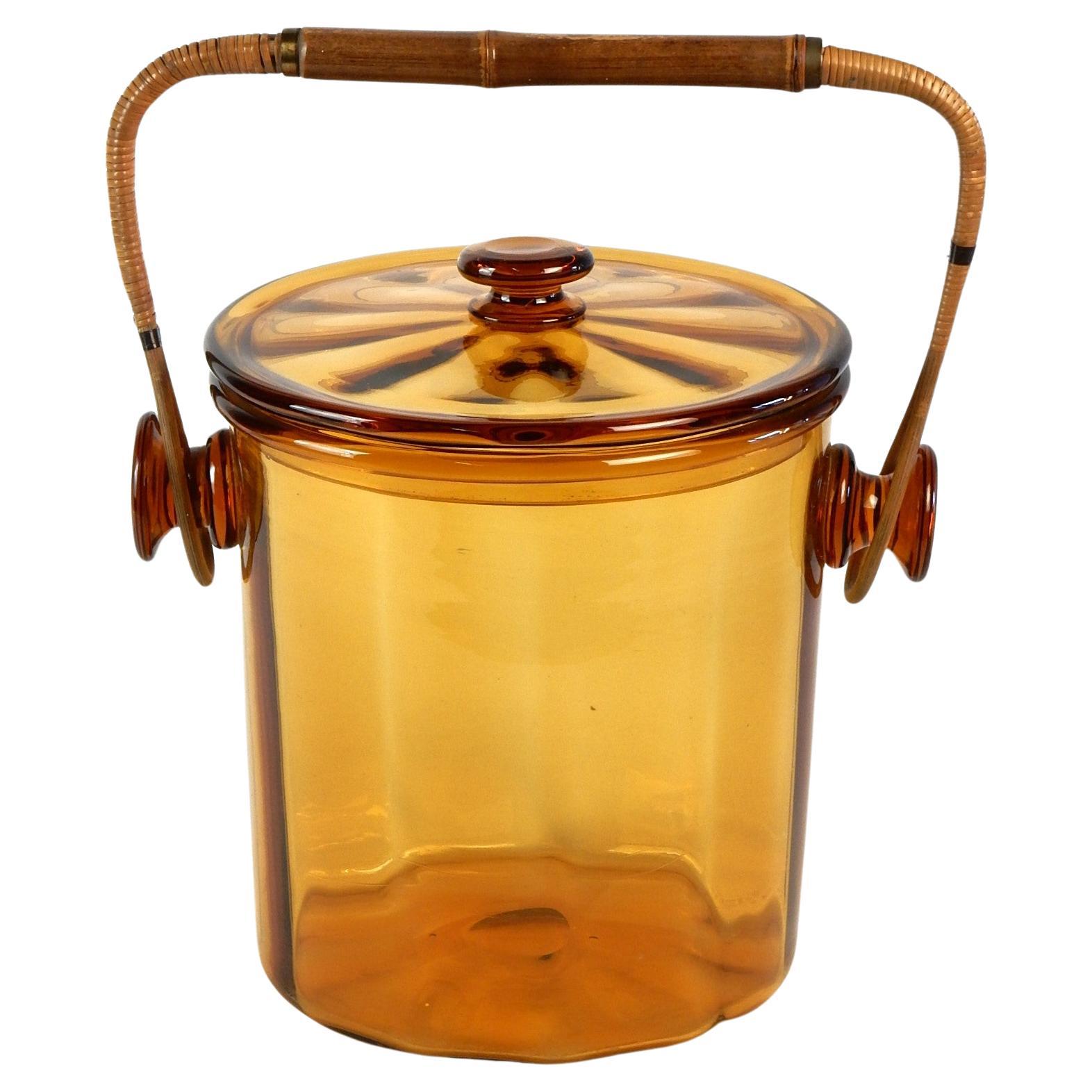 Exquisite art glass lidded ice bucket with rattan handle wrapped in cane.
Gorgeous honey colored glass. Large piece measuring 12in X 11in (inside 9-1/2in tall X 8.50in wide). 
Not signed. From a modernist 1960's estate with many Austrian and French