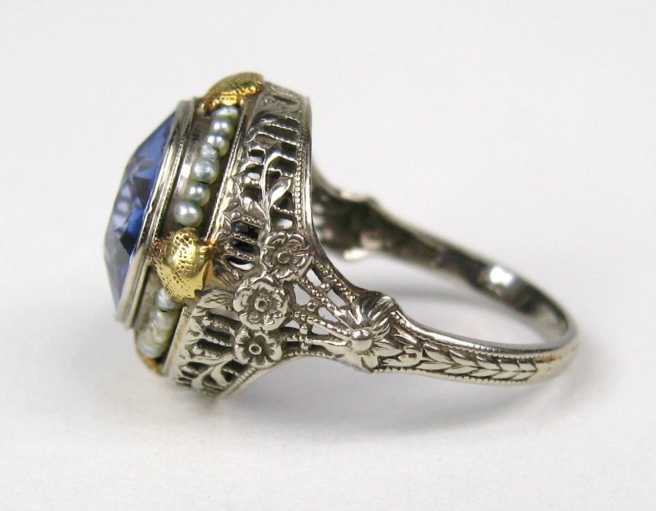 Set in 14K White gold with yellow gold accents. Bezel Set stunning Synthetic Blue Sapphire with tiny seed pearls surrounding Floral motif on the shank. The Ring is a size 7-3/4 and can be sized by us or your jeweler. This is out of a massive