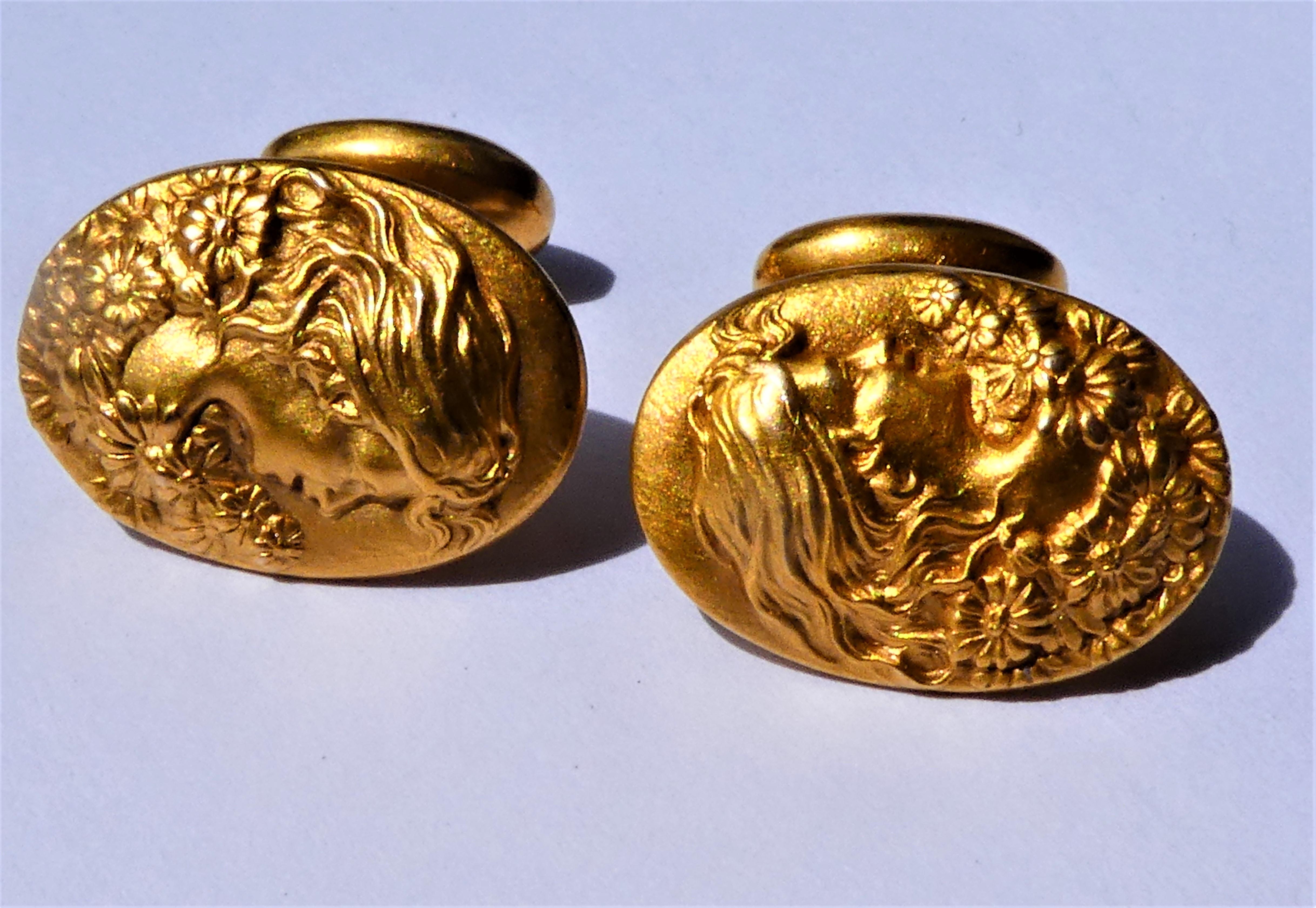 The oval cufflinks show a smiling lady with long hair and flowers. They were crafted around 1900 by an American maker in 14 karat yellow gold and are slightly curved on the edge. The solid gold cuff buttons have a dumb-bell link. They are