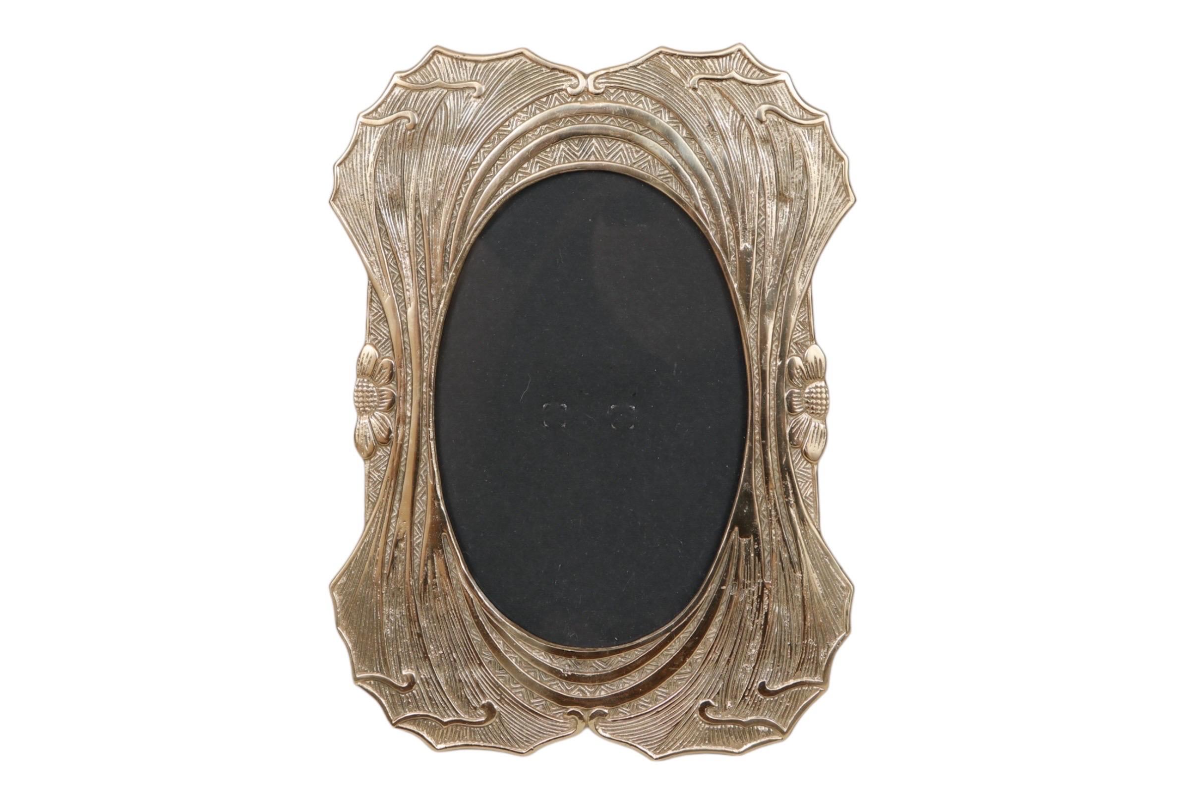 An Art Nouveau 5” x 7” pressed brass photo frame. The oval picture window is framed with an elegant flowing design of undulating lines resembling the stems of plants. Each side is decorated with a simple flower. Plant like formations are