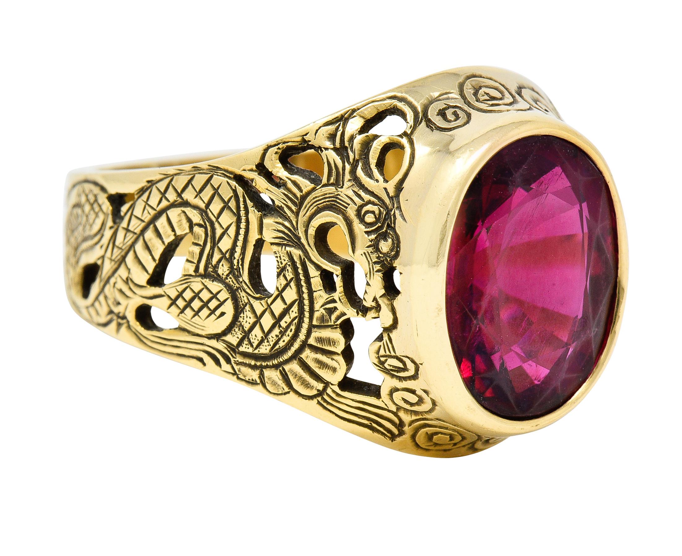 Centering an oval cut rubellite weighing approximately 7.85 carats total. Transparent medium purplish red in color and bezel set. Flanked by ornate pierced dragon motif shoulders. Engraved with texturous scales, tail, and smoke. Stamped 18k for 18