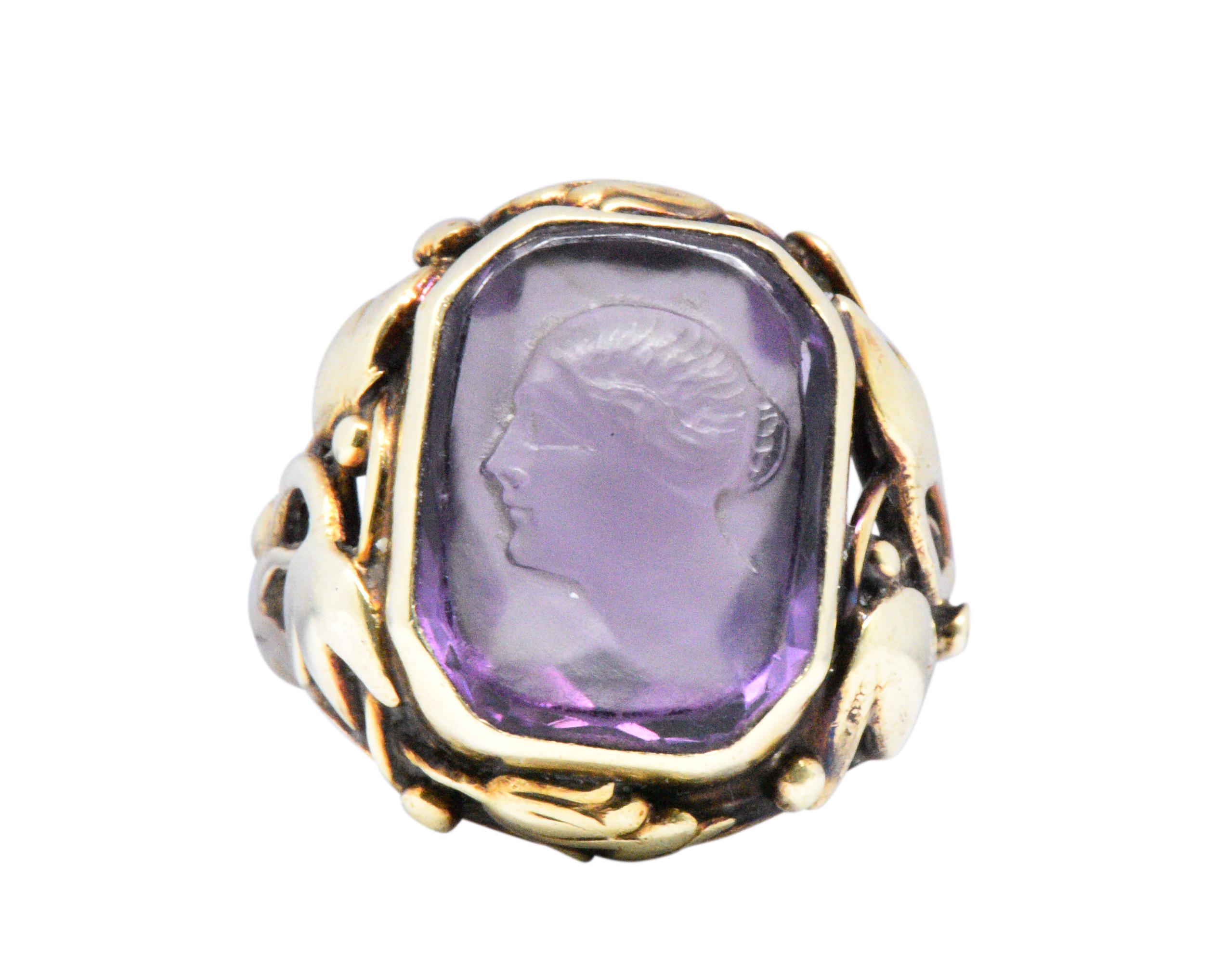 Centering a cut-corner amethyst measuring 15.5 x 11.6 mm, carved depicting the profile of a woman, facing left, wonderfully detailed, you can see her hair combed into a bun

Bezel set with a foliate motif setting, in lovely green gold, wrapping