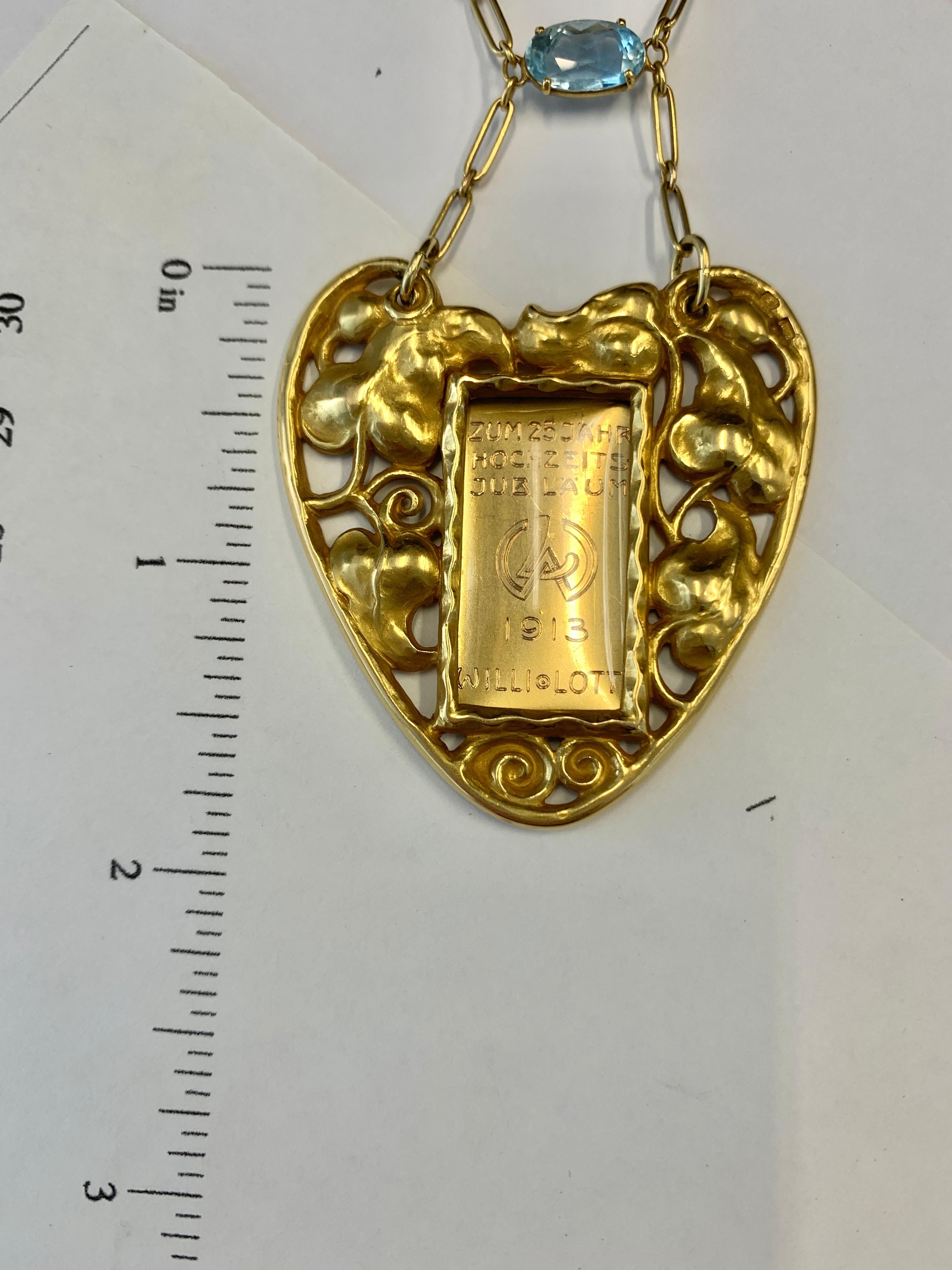 !8kt Gold heart-form necklace pendant with scrolling leaf and vine motifs, centering a glass picture frame with an oval-cut aquamarine (weighing approx. 1.30cts) accent. Engraved on the back with date and inscription. Austro-Hungarian maker's mark