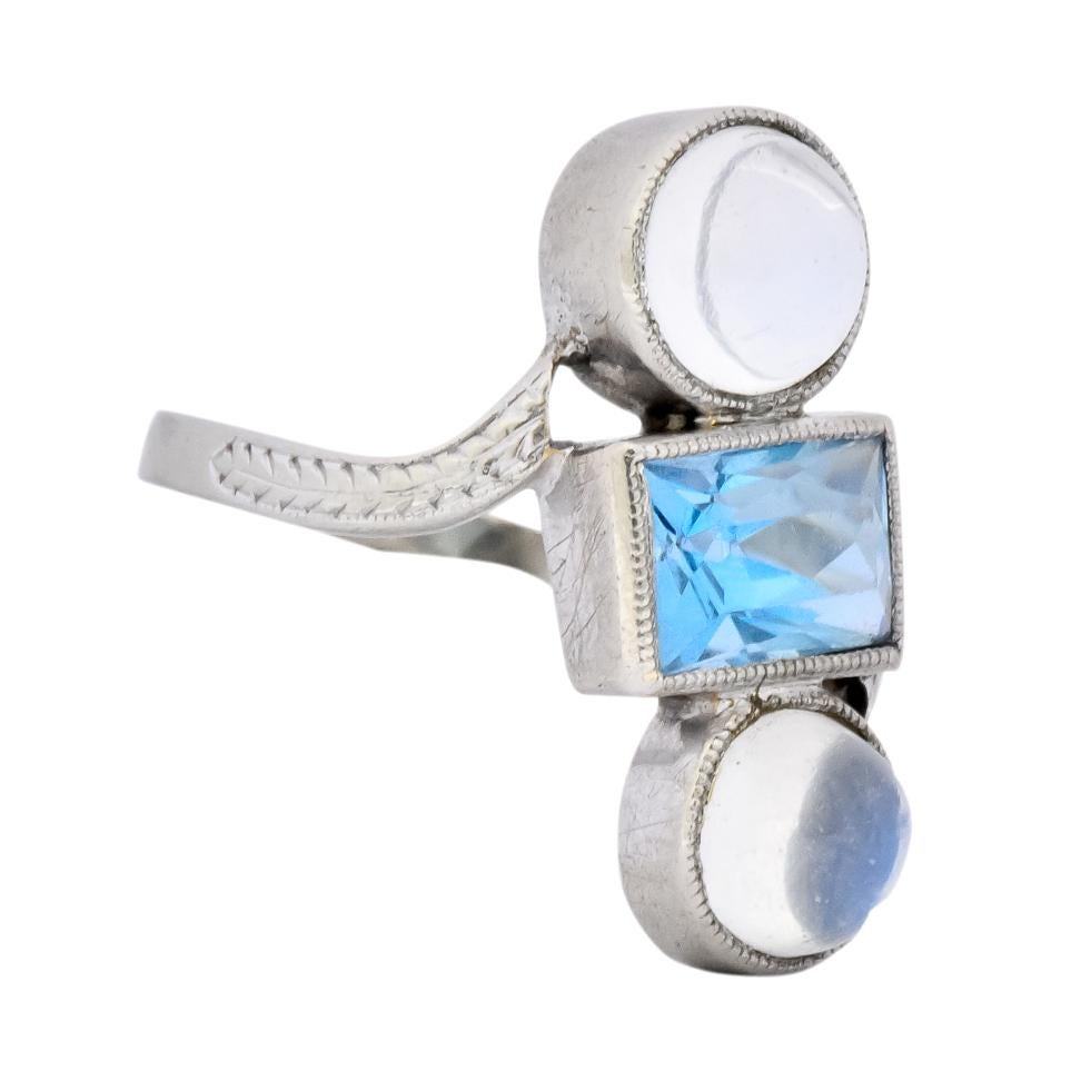 Centering a scissor cut aquamarine measuring approximately 7.0 x 5.5 mm, transparent and a medium sky blue color

Flanked by two round cabochon cut moonstones measuring approximately 6.0 mm, transparent with moderate blue adularescence

Bezel set