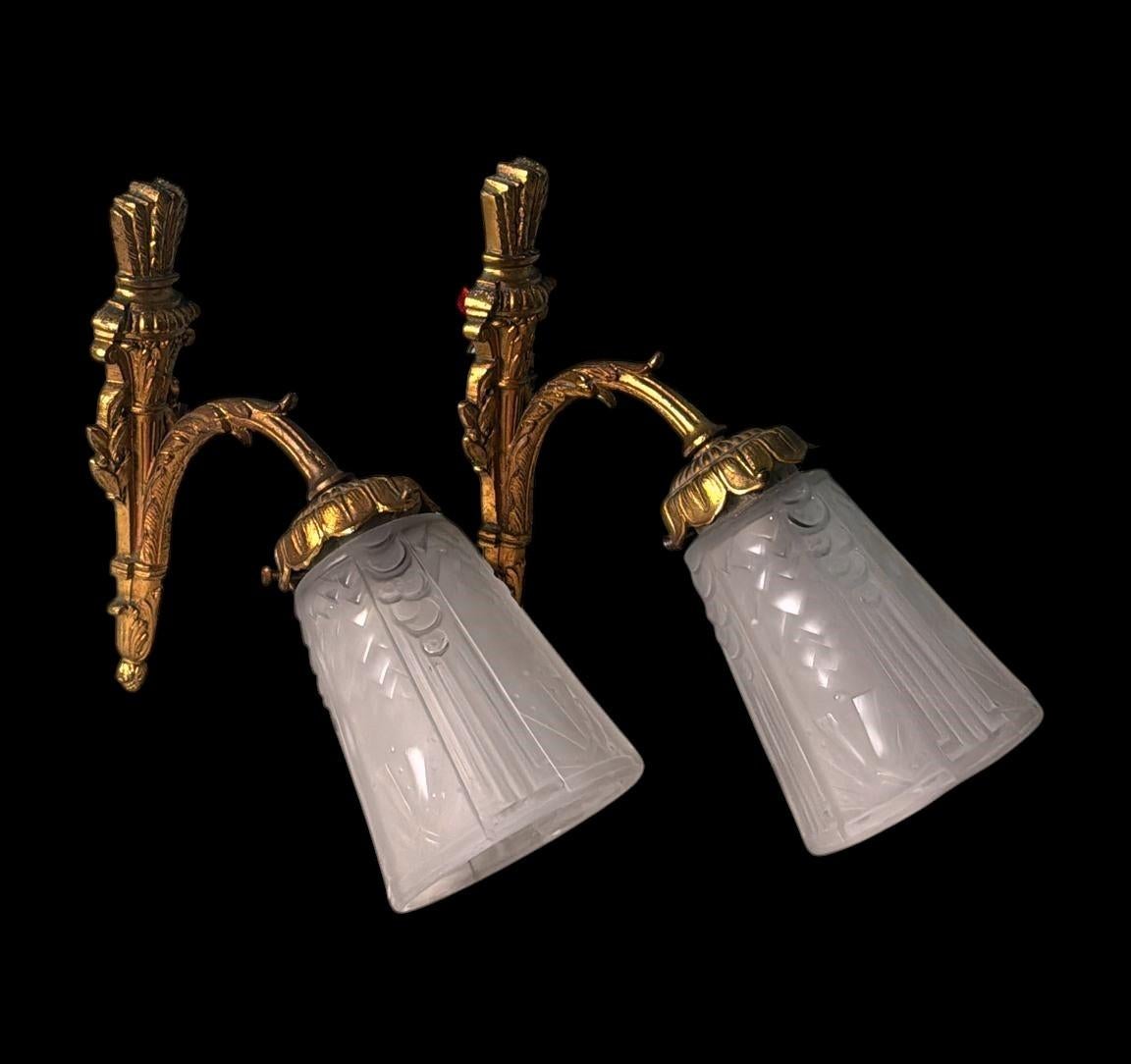 Questa raffinata coppia di applique francesi incarna lo spirito elegante della Parigi degli anni Venti. Le montature in bronzo dorato, finemente fuse e ricche di dettagli con fogliame e motivi floreali, sostengono paralumi in vetro tagliato su