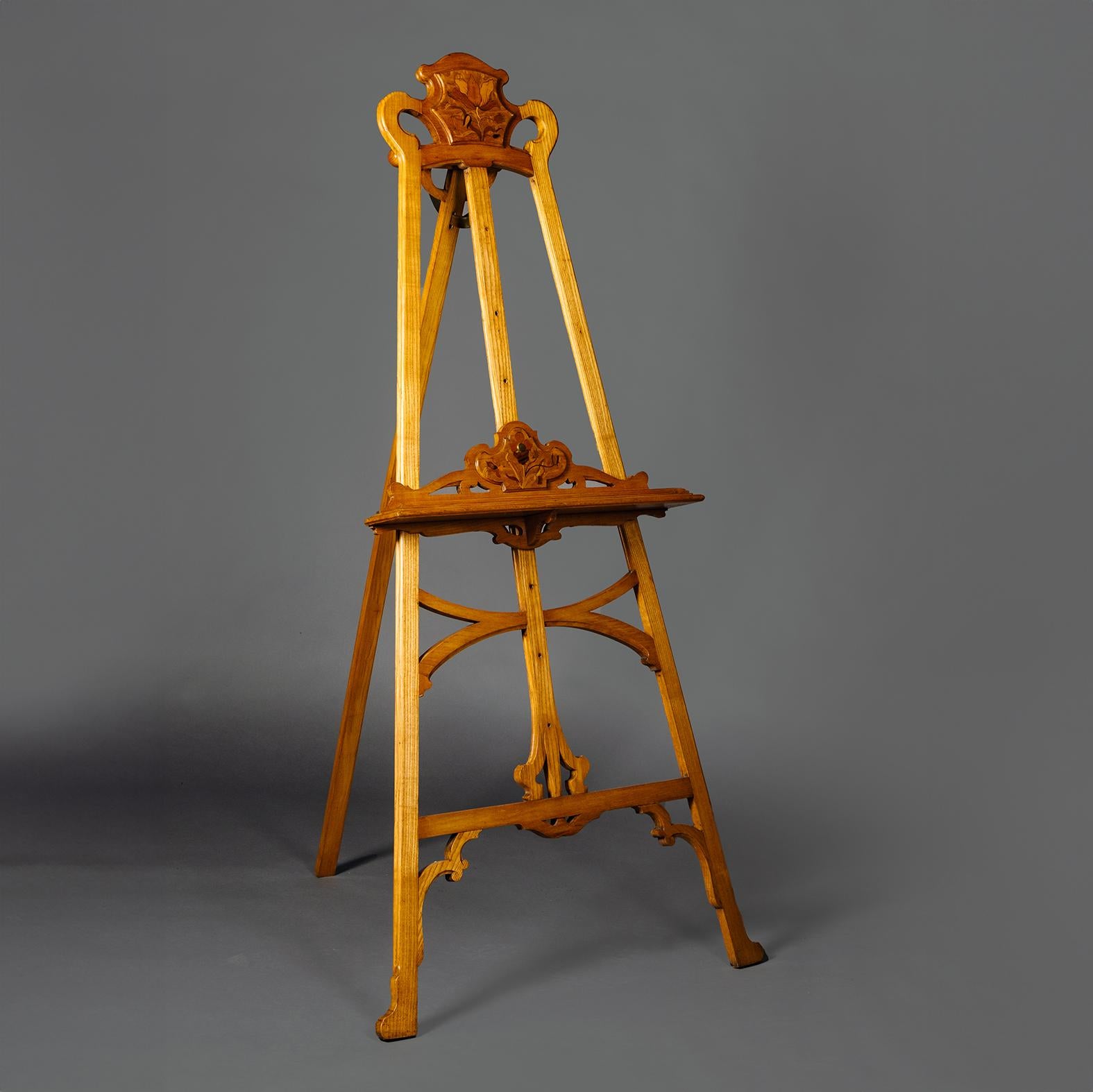 An Art Nouveau Ash and Marquetry Easel. 

The shaped cresting with marquetry panel of coloured woods depicting a Lilly flower, above a splayed latticework stand with an adjustable rack and support decorated with marquetry flowers.

This elegant