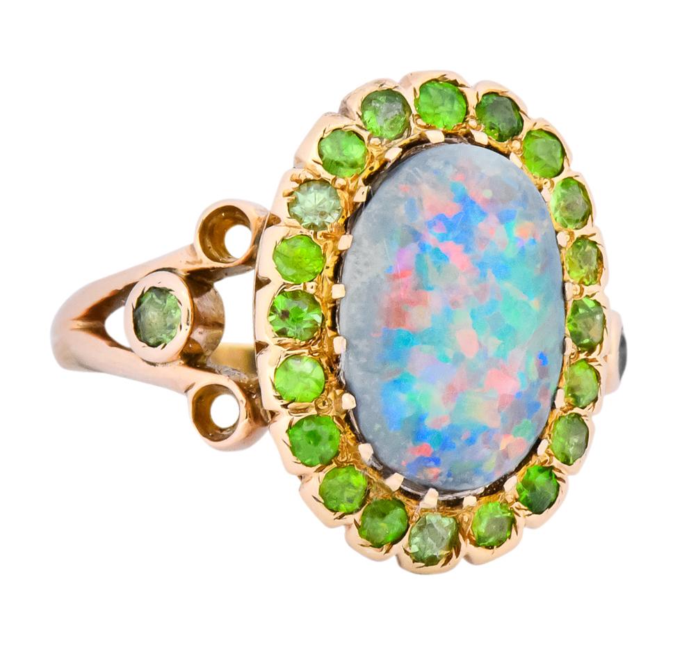 Cluster style ring centering an oval cabochon cut black opal measuring approximately 12.3 x 8.3 mm, excellent play of color with strong flashes of spectral color

Surrounded by single and Swiss cut demantoid garnets (worn) weighing approximately
