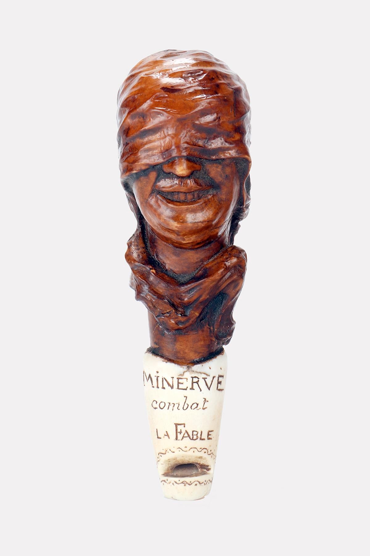Un fischietto Art Carved in legno di bosso intagliato, raffigurante il volto sorridente di una donna bendata. La parte terminale, il fischietto vero e proprio, è in corno di cervo, con incisa la frase in francese: Minerva combat la Fable (Minerva