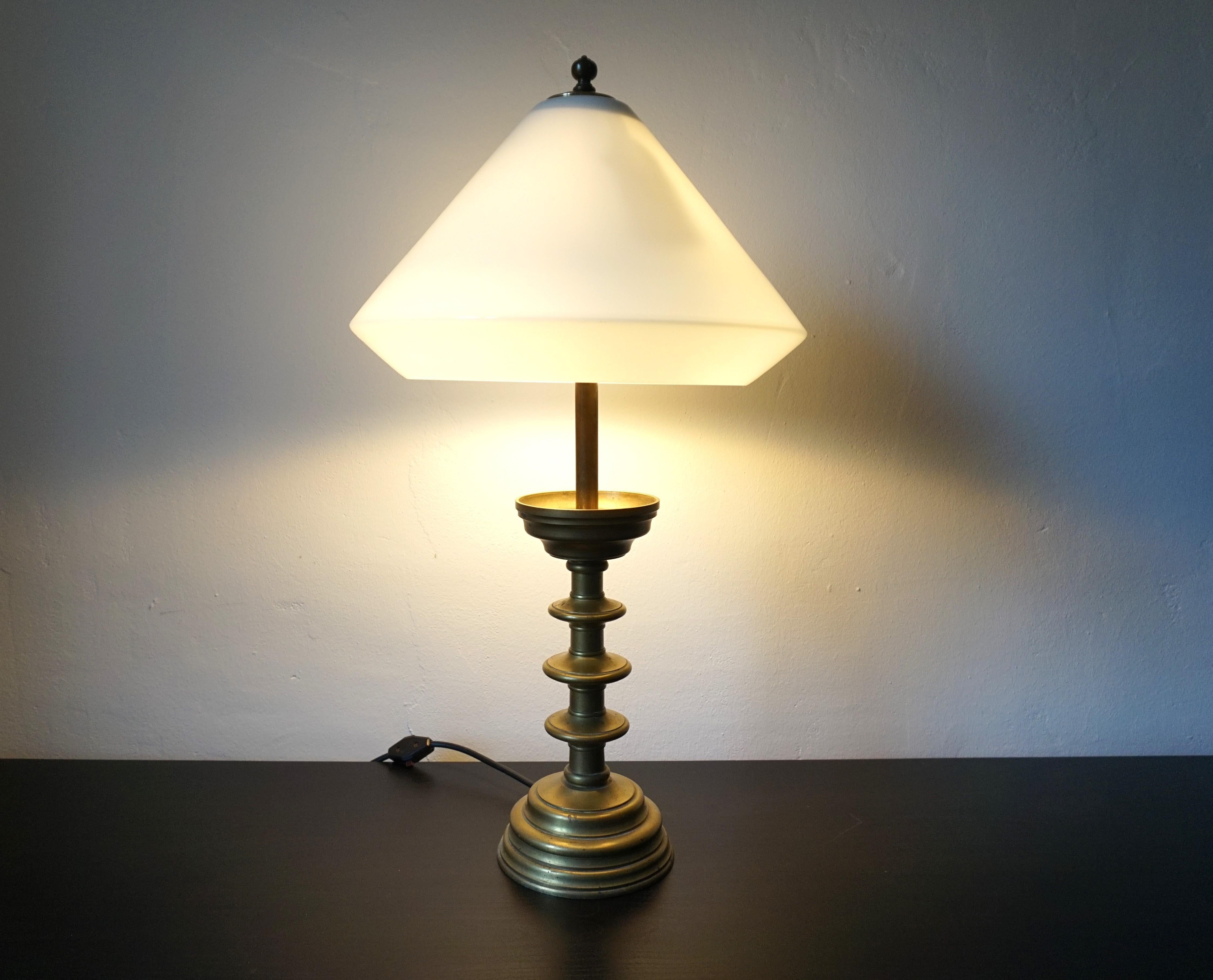 This well proportioned solid brass table lamp comes in excellent condition with a fantastic patinated lamp base.  The foot consists of several plate-like elements and is formed into a bowl at the top. The cone-like lampshade made of light opal glass