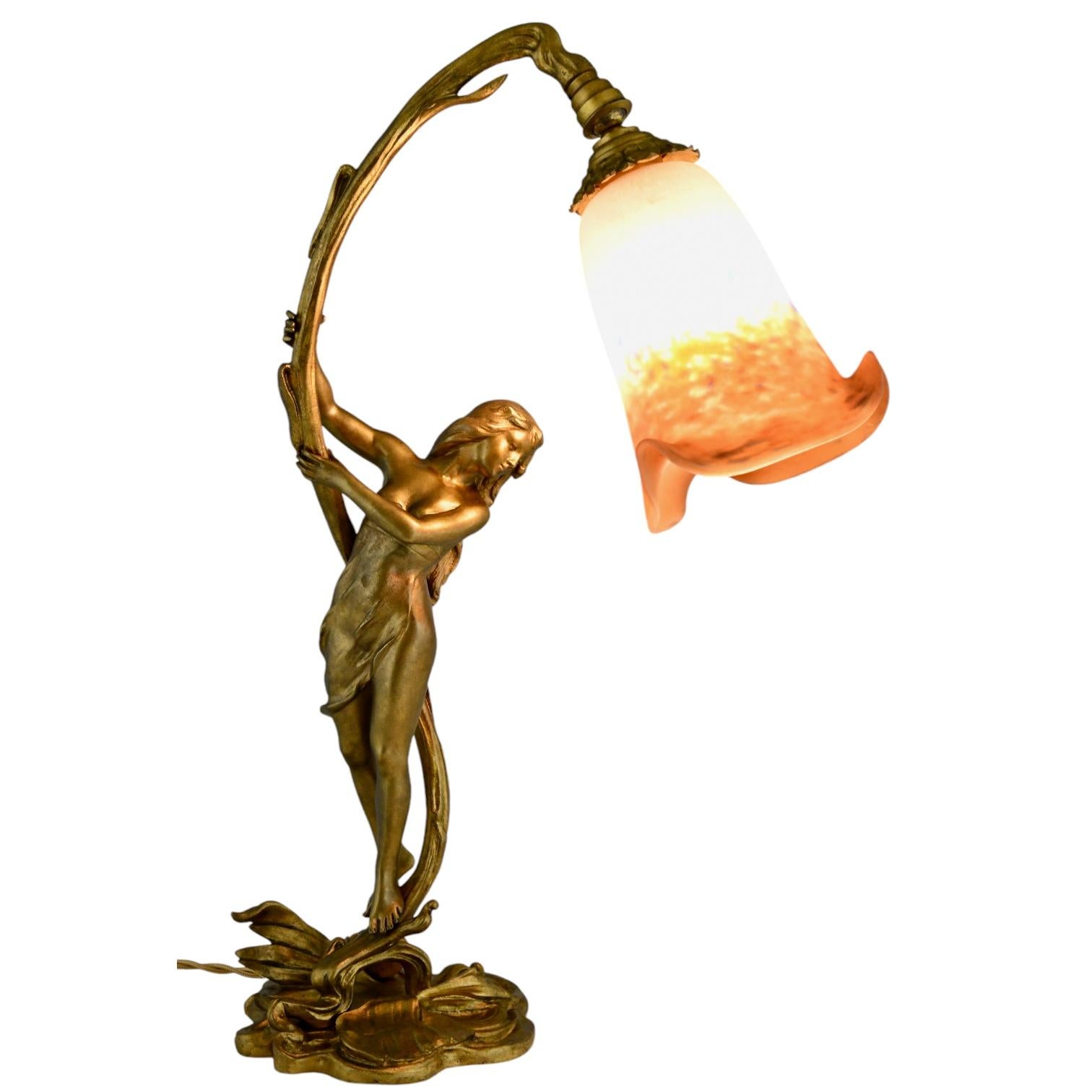 Lámpara Art Nouveau de bronce con dama de Ferville Suan, bronce patinado con pantalla de cristal firmada Degué. 
Firma fundadora Lebrun.
Pantalla de cristal pate de verre naranja, azul y blanco. 
Francia ca. 1900. 
Literatura:
Etains 1900, Philippe