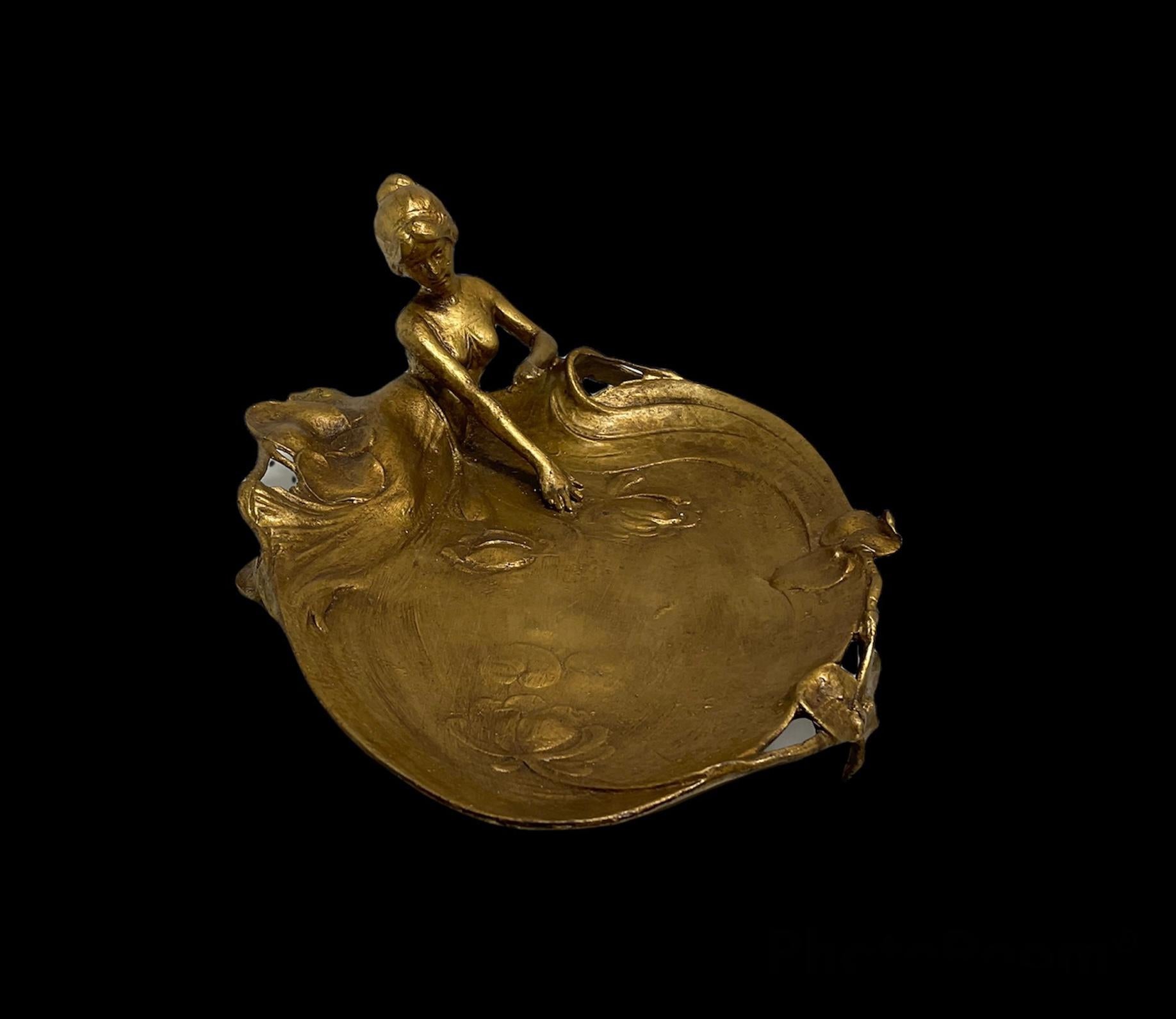 This is an Art Nouveau bronzed Metal jewelry, pin, coin tray or soap dish. It is depicting a maid with a long dress laying down at the edge of a lily pond. She is trying to get hold of one of the flowers that are floating. An unrecognized mark below