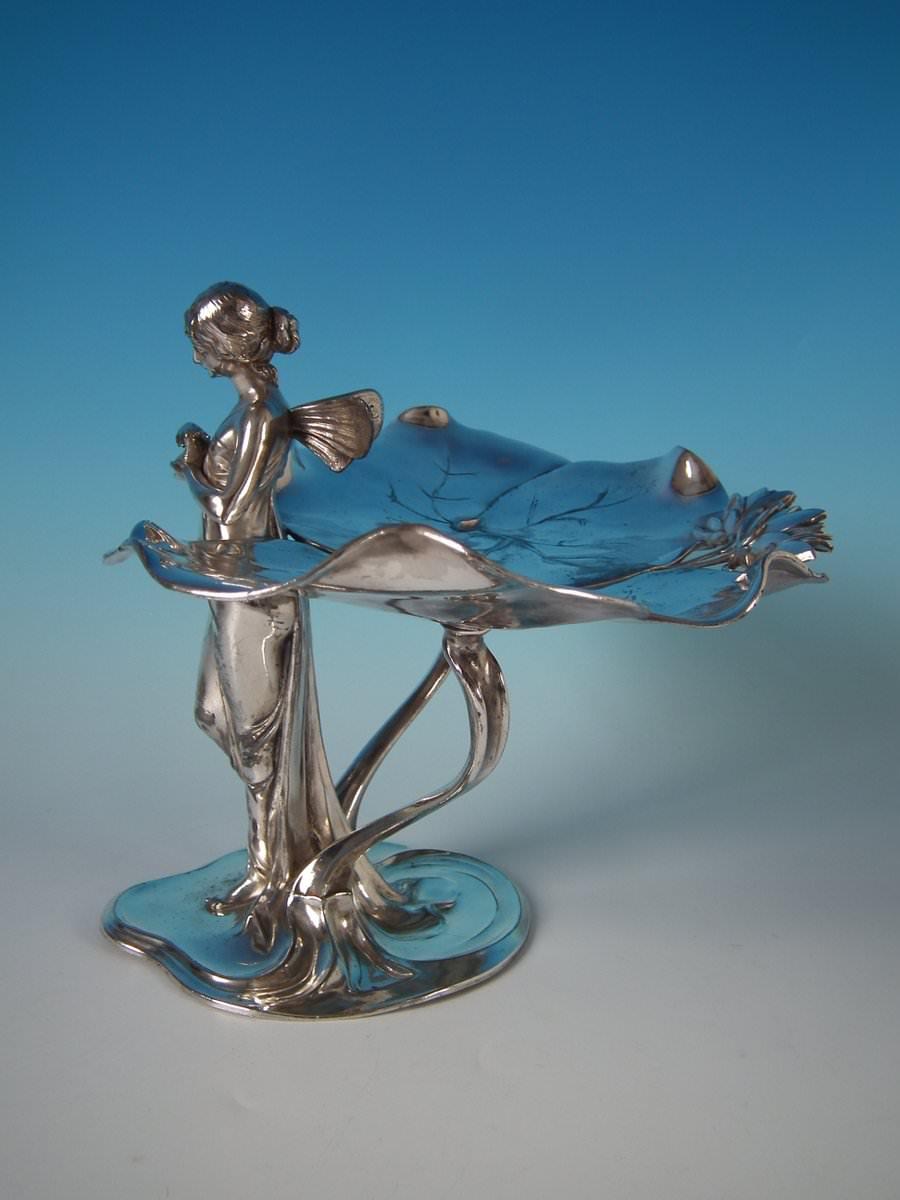 WMF Art Nouveau visiting card tray decorated with a fairy standing before two large lily leaves, holding a dove in her hands. Impressed maker's marks including WMF, 'E', 'I/O', and '246' to the underside. Illustrated on page 167, catalogue number