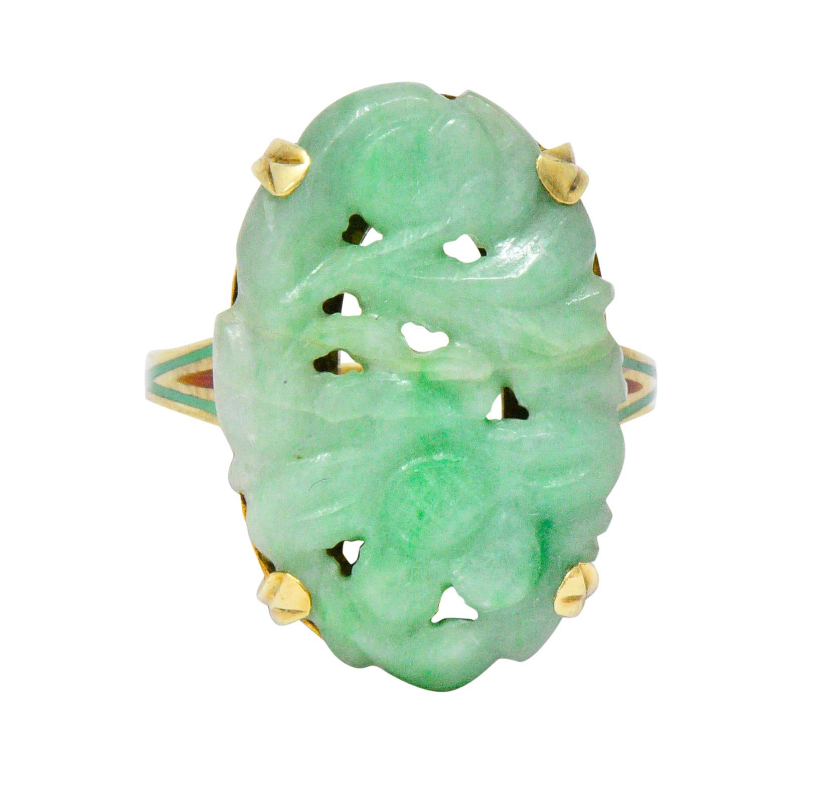 Centering an oval carved jade, measuring approximately 22.5 x 15.2 mm, light mottled green

Carved with a floral and foliate motif

Shoulders exhibit inlaid green and deep orange enamel in a chevron motif

With maker's mark and stamped 14K

Ring