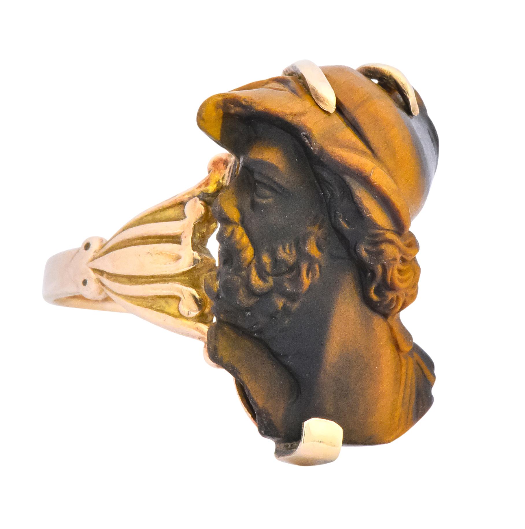 Centering a hand carved tiger's eye stone depicting a Greek hero with helmet and stylized curling beard

Measuring 3/4 x 1/2 inch, opaque with dark to light brown banding and slight chatoyancy

Claw set and flanked by two gold lotus flower motifs on