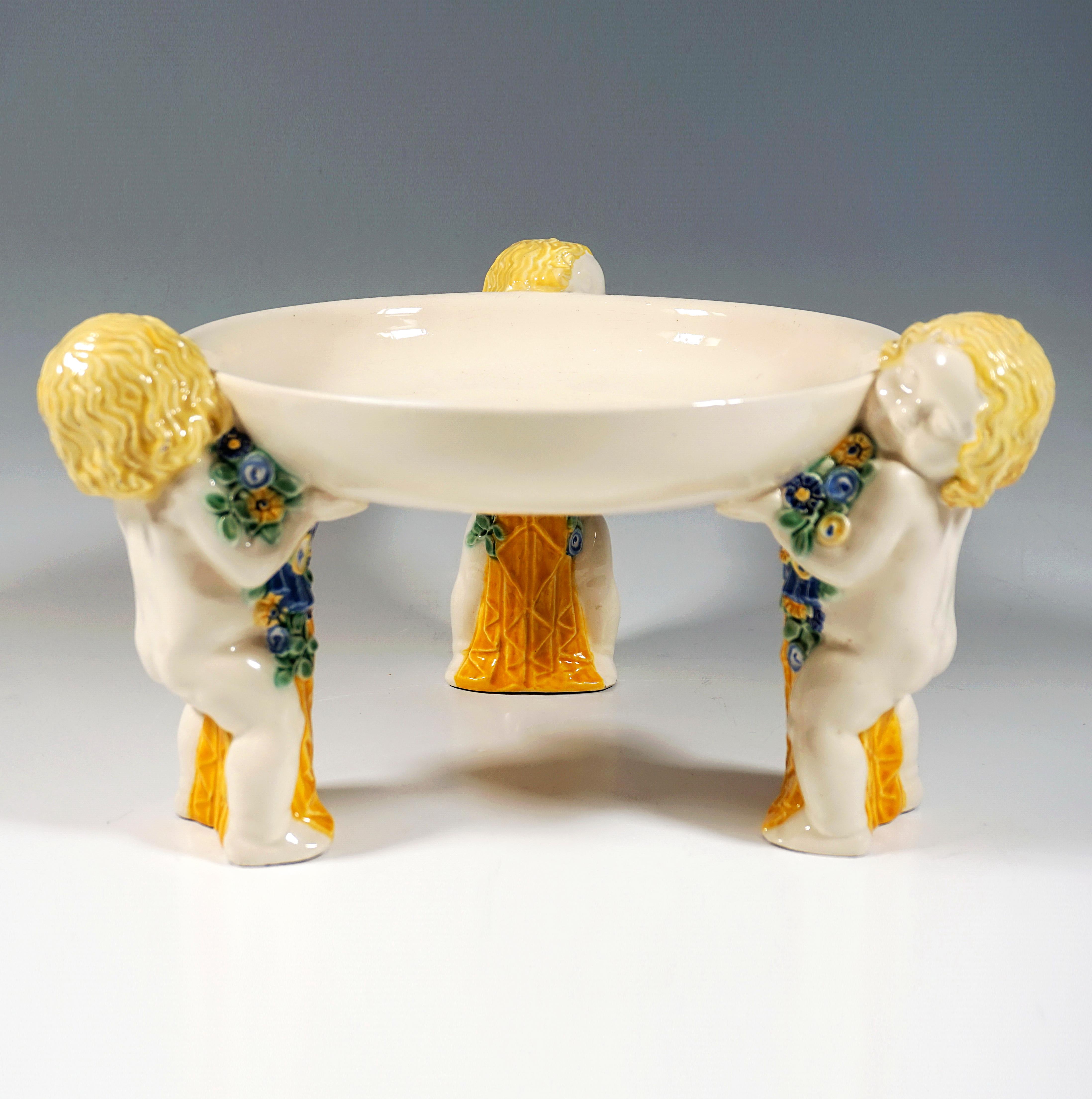 Three standing nude putti with floral decorations and yellow draperies carrying a shallow round bowl. Without base.

By Michael Powolny (1871 - 1954)
Nowadays, art experts attest Michael Powolny’s work as the work of one of the most important