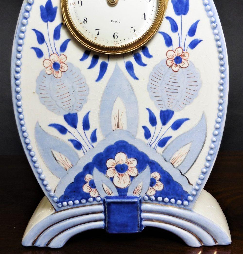 Art Nouveau mantel clock in a ceramic case with a white ground decorated with flowers in shades of blue with beaded decoration to the outside of the case, standing on a ribbed outswept arch with rectangular centre.
Gilded bezel opening to an enamel