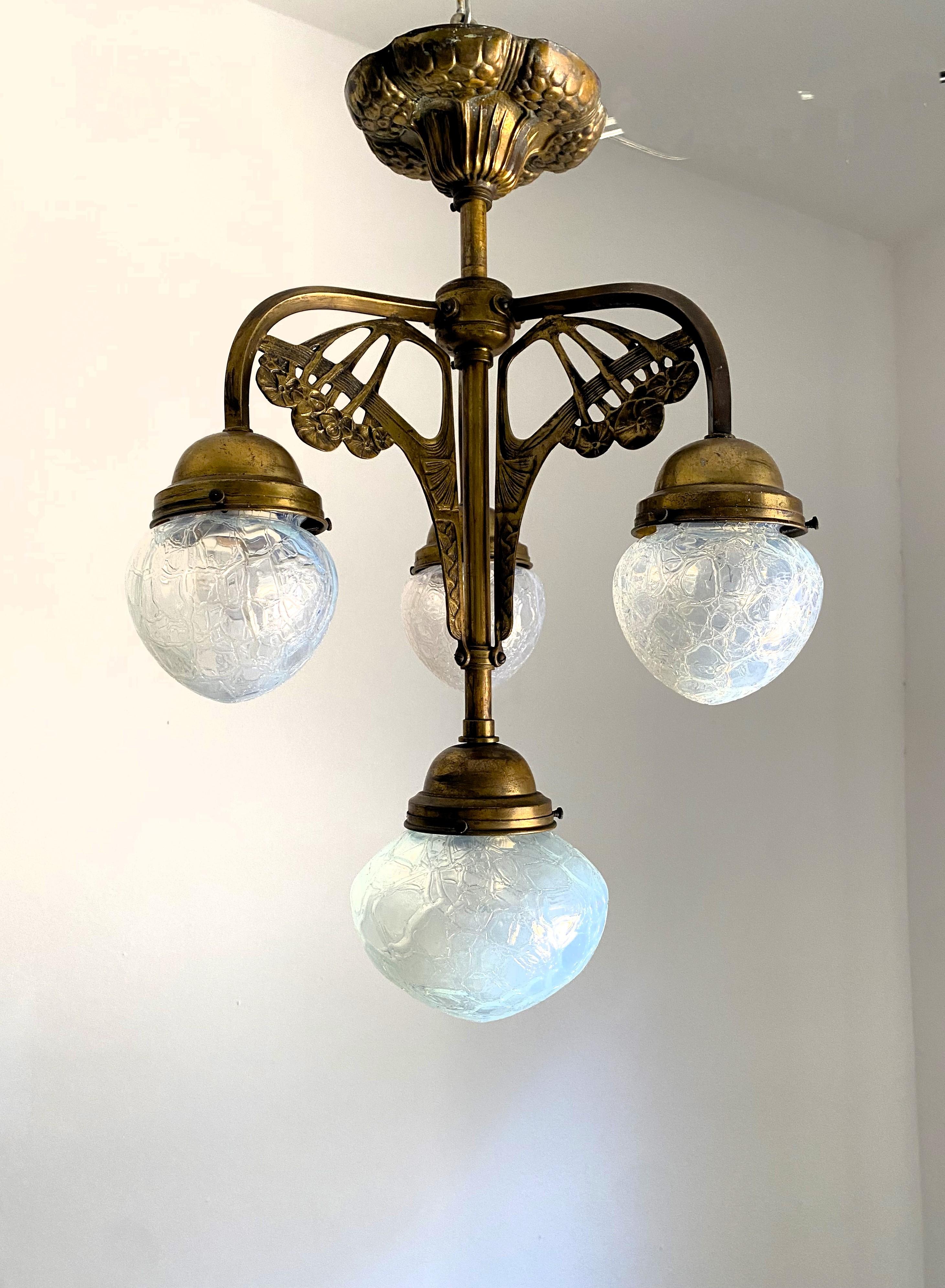 Art Nouveau chandelier manufactured in brass and hand blown opalescent glass.
The structure of the chandelier is adorned by floral motifs and shows a dark patina throughout, this can be polished back to golden easily if the client wants us to do