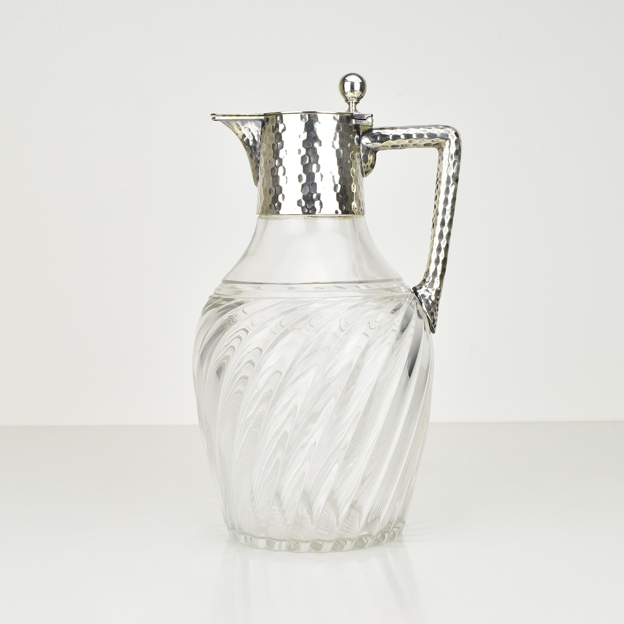 Este exquisito decantador de cristal tallado Art Nouveau, también conocido como jarra de clarete, presenta un montaje de plata martillada a mano de impresionante detalle, obra de Wilhelm Theodor Binder. El cuerpo de cristal presenta un elegante
