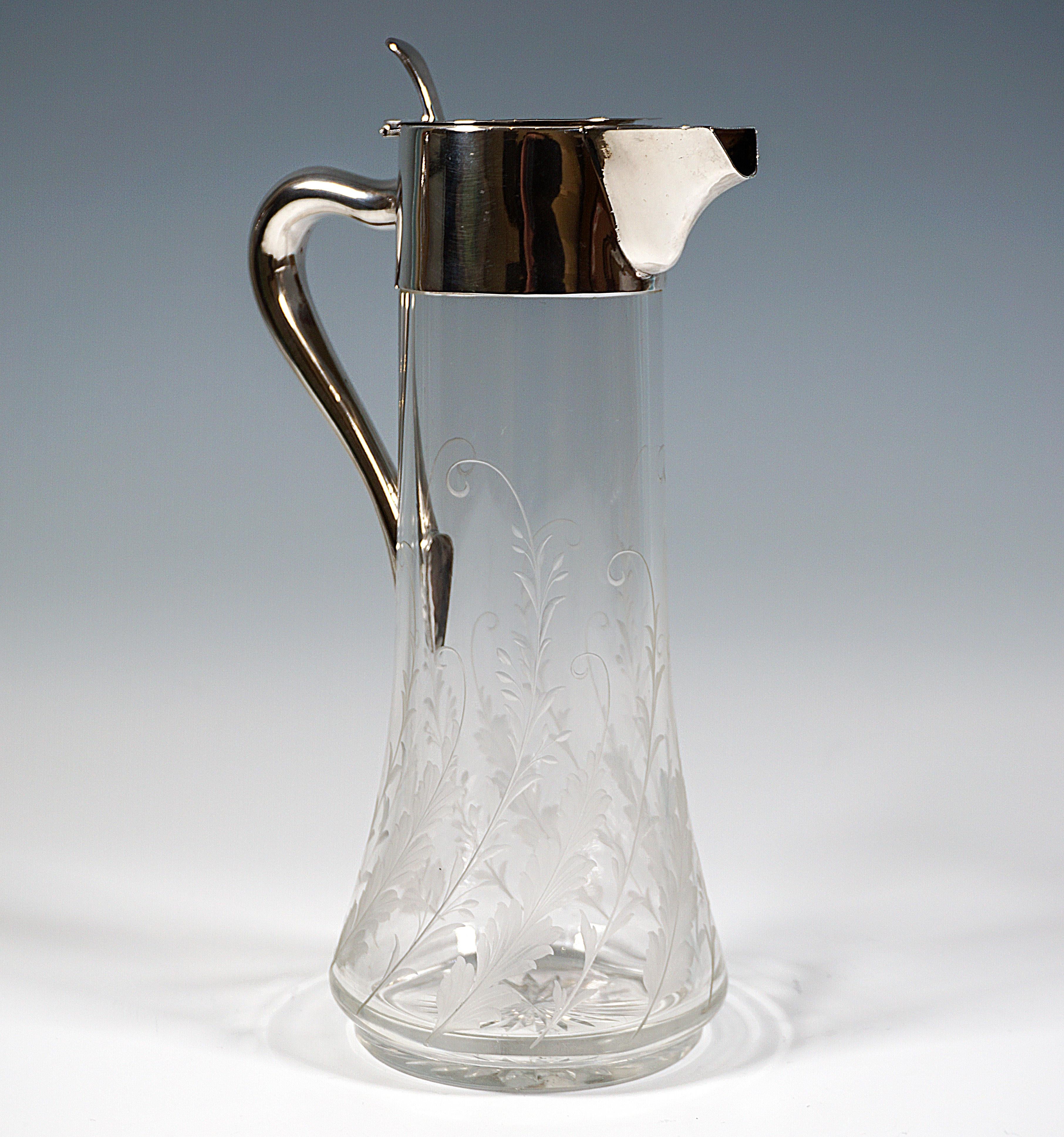 Jarra Art Nouveau de vidrio transparente con cuerpo cónico, zarcillos de hojas de pluma finamente tallados que suben en espiral desde el borde inferior, estrella recortada esmerilada, montura lisa de plata con asa curva en forma de J, pitorro