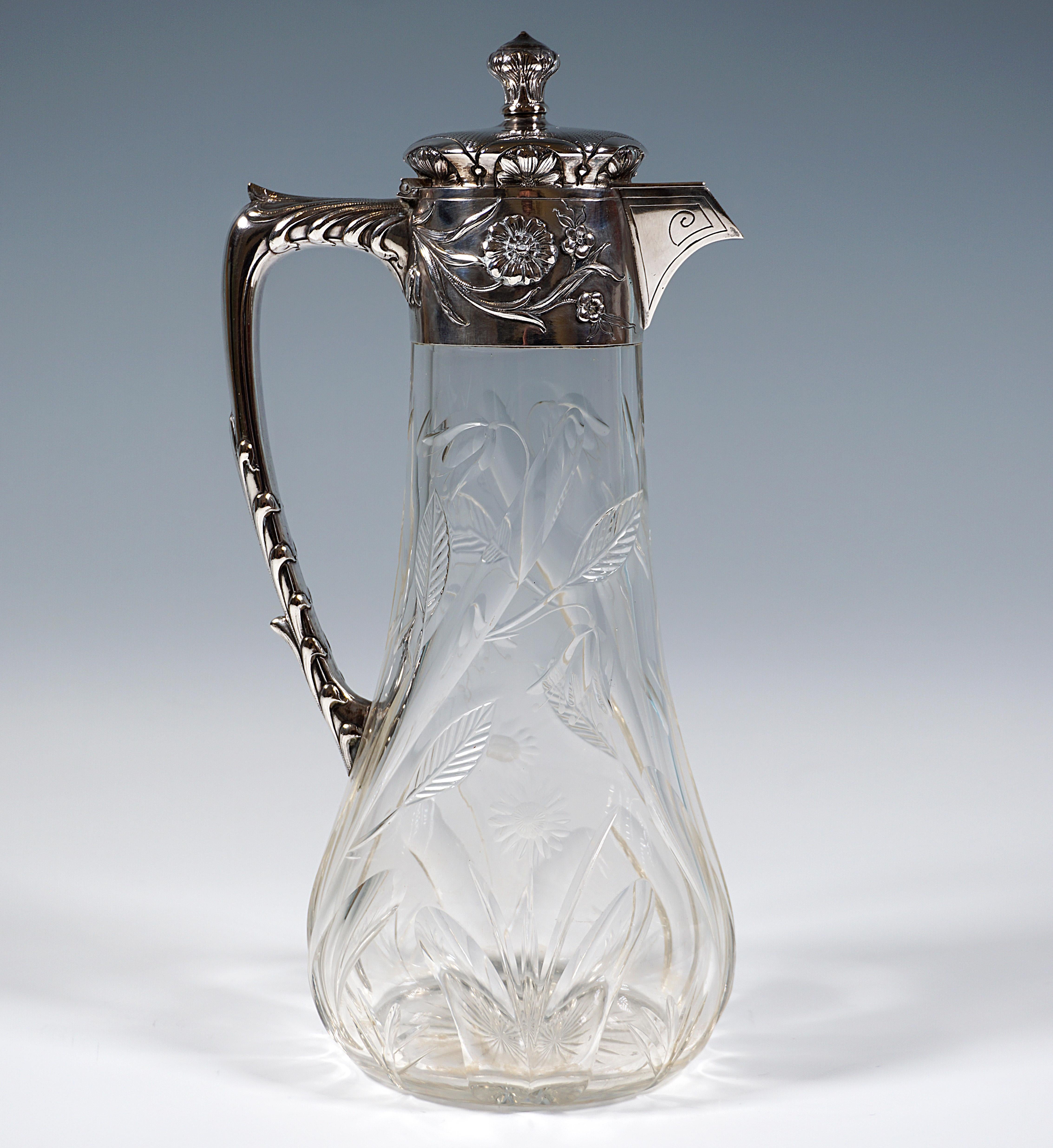 Art Nouveau carafe made of clear glass with a conical body on an oval base, cut meadow flower decoration rising from the lower rim, cut ground star, silver mount decorated with floral relief decoration with a J-shaped handle, covered, beak-shaped