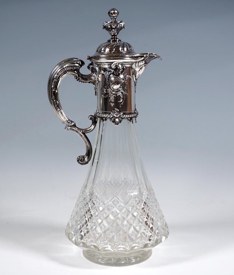 Carafe made of clear glass with a conical, faceted body that swings out at the bottom with a separate base, the lower half has a stone cut decoration with stars cut in the middle and a star on the bottom, silver fittings with rocaille decoration and