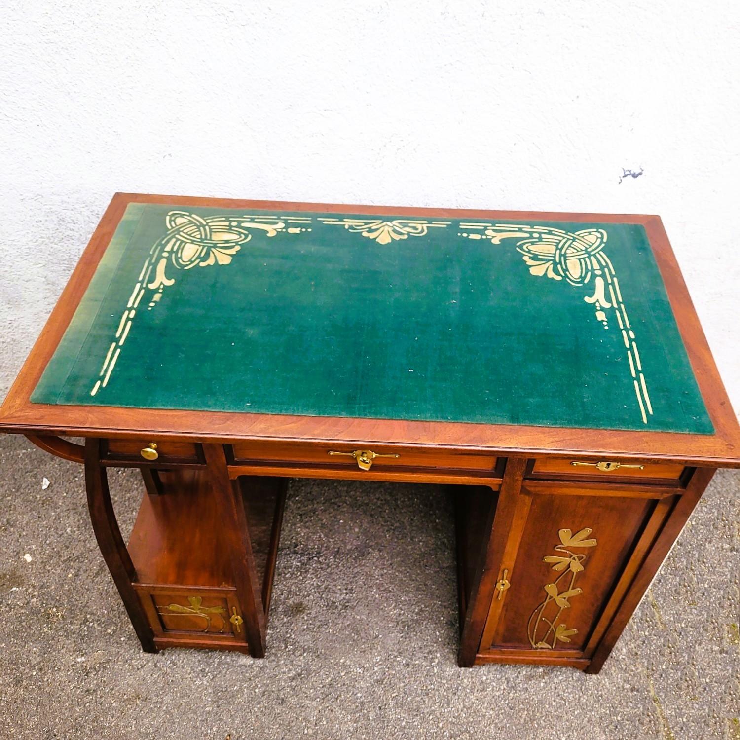 Beautiful Art Nouveau wooden desk with rich floral decoration in brass cut and fixed to the wood.  

 It opens with 3 drawers and 2 doors, the bigger one contains sliding drawers.   

The top is covered with a green fabric and a glass top.   

Good