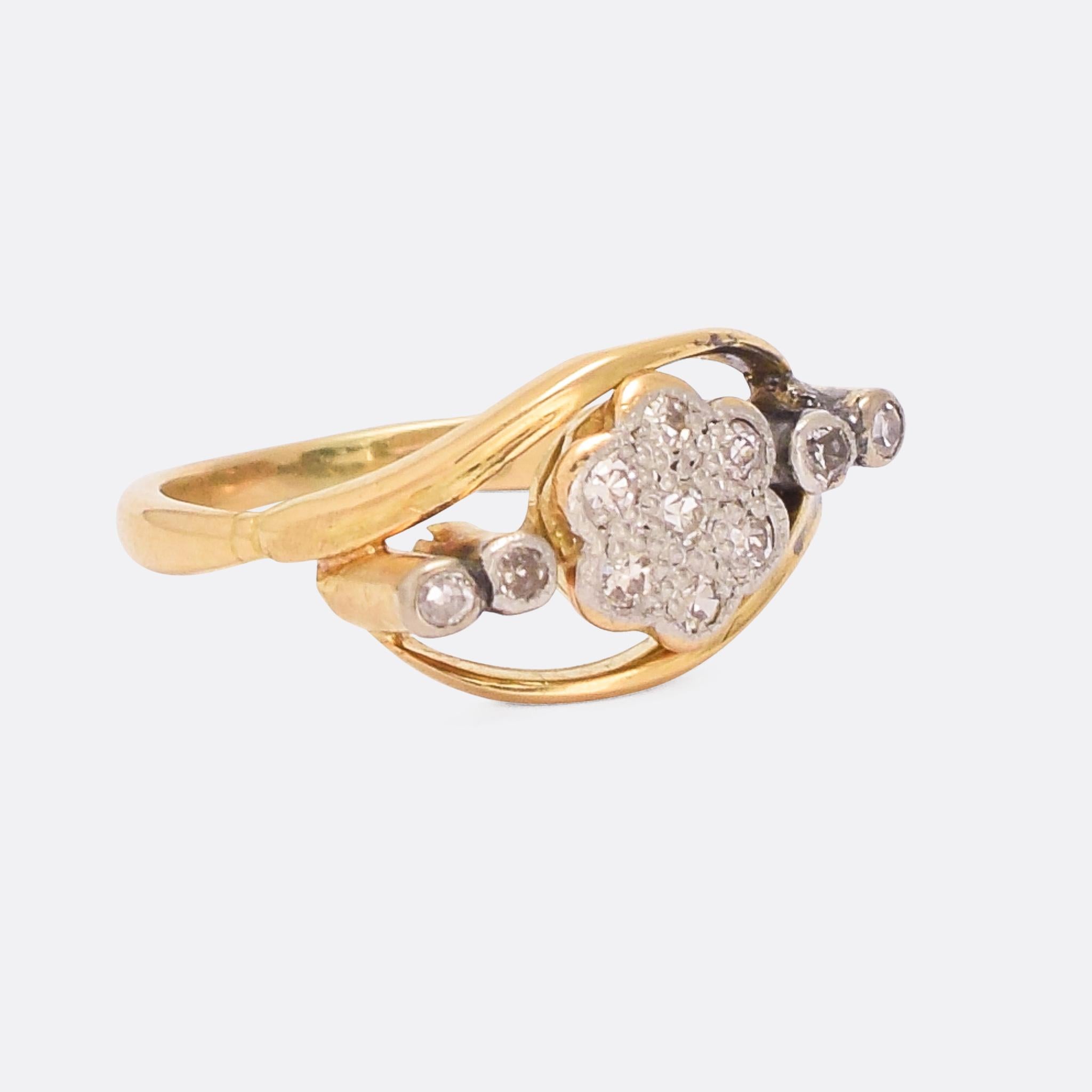 A sweet Art Nouveau crossover ring dating from the early 20th Century. The head features a diamond daisy cluster, with two diamond accents to either side and flowing shoulders that continue all the way around. It's crafted in 18 karat gold and