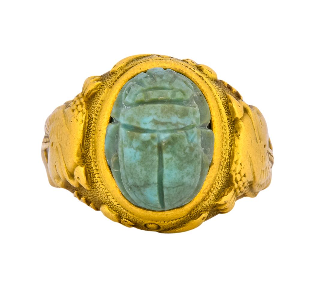 Centering a deeply carved turquoise scarab measuring 11.1 x 8.3 mm, opaque and greenish-blue in color with brown matrix veining

Bezel set into matte gold mounting with a stippled texture and hieroglyphic motif

Each shoulder depicts an Egyptian