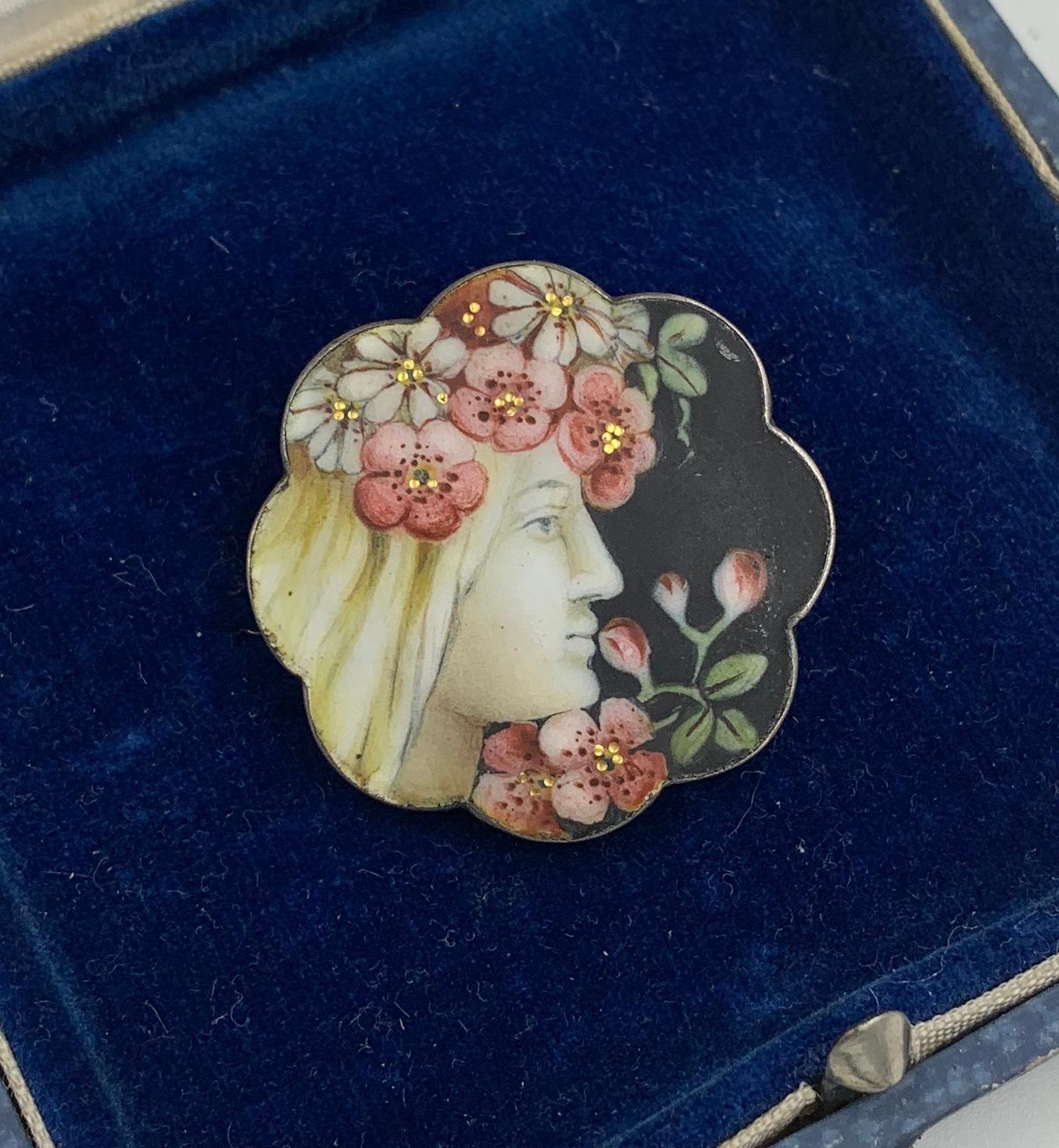 Une très rare broche Art Nouveau de qualité musée en émail peint à la main sur de l'argent sterling avec des reflets dorés.   Une belle jeune fille est représentée avec des fleurs roses et blanches dans les cheveux, de jolies feuilles vertes et des