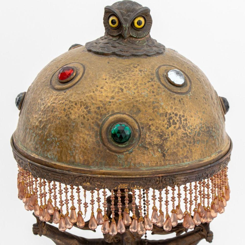 Lampe figurative Art Nouveau en étain doré et patiné, l'abat-jour en métal décoré de cabochons en verre multicolore facettés et surmonté d'un fleuron en forme de hibou, avec une frange perlée. 22