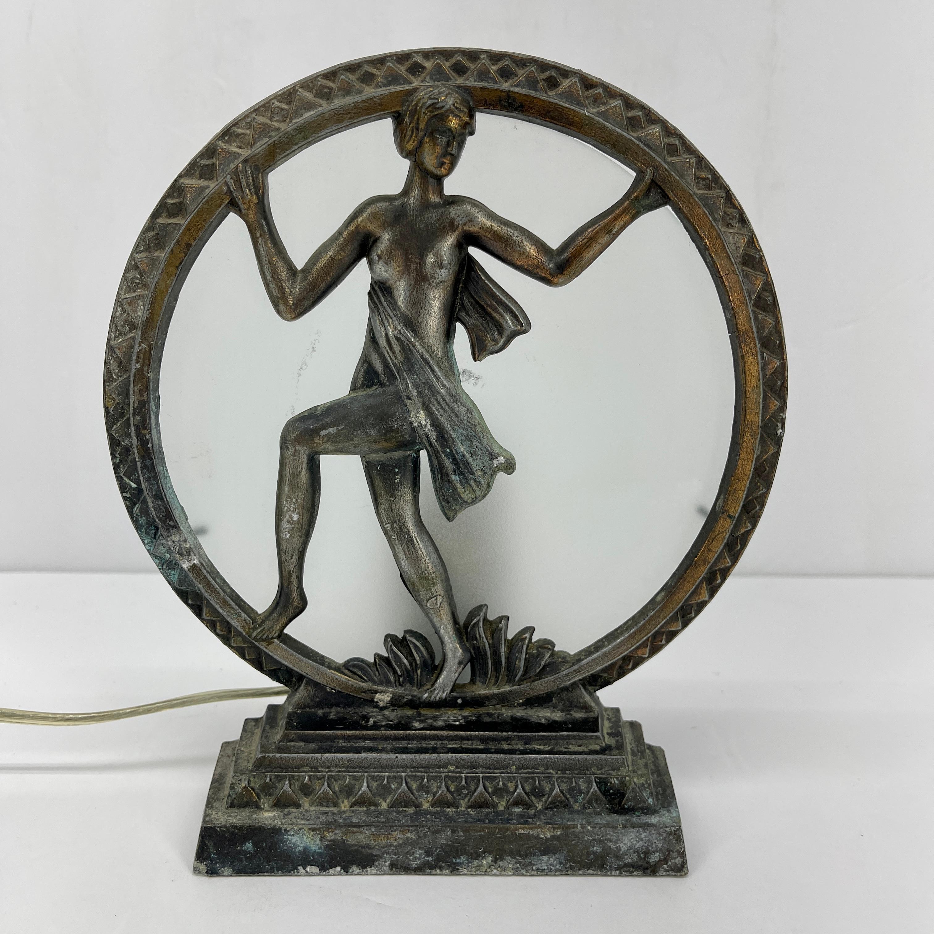 Art Nouveau figural woman table lamp. This beautiful petite lamp is a classic piece of Art Nouveau design. The nude woman graces with elegant the front of the lamp with circular glass behind her. The light shines from the back enhancing her beauty.