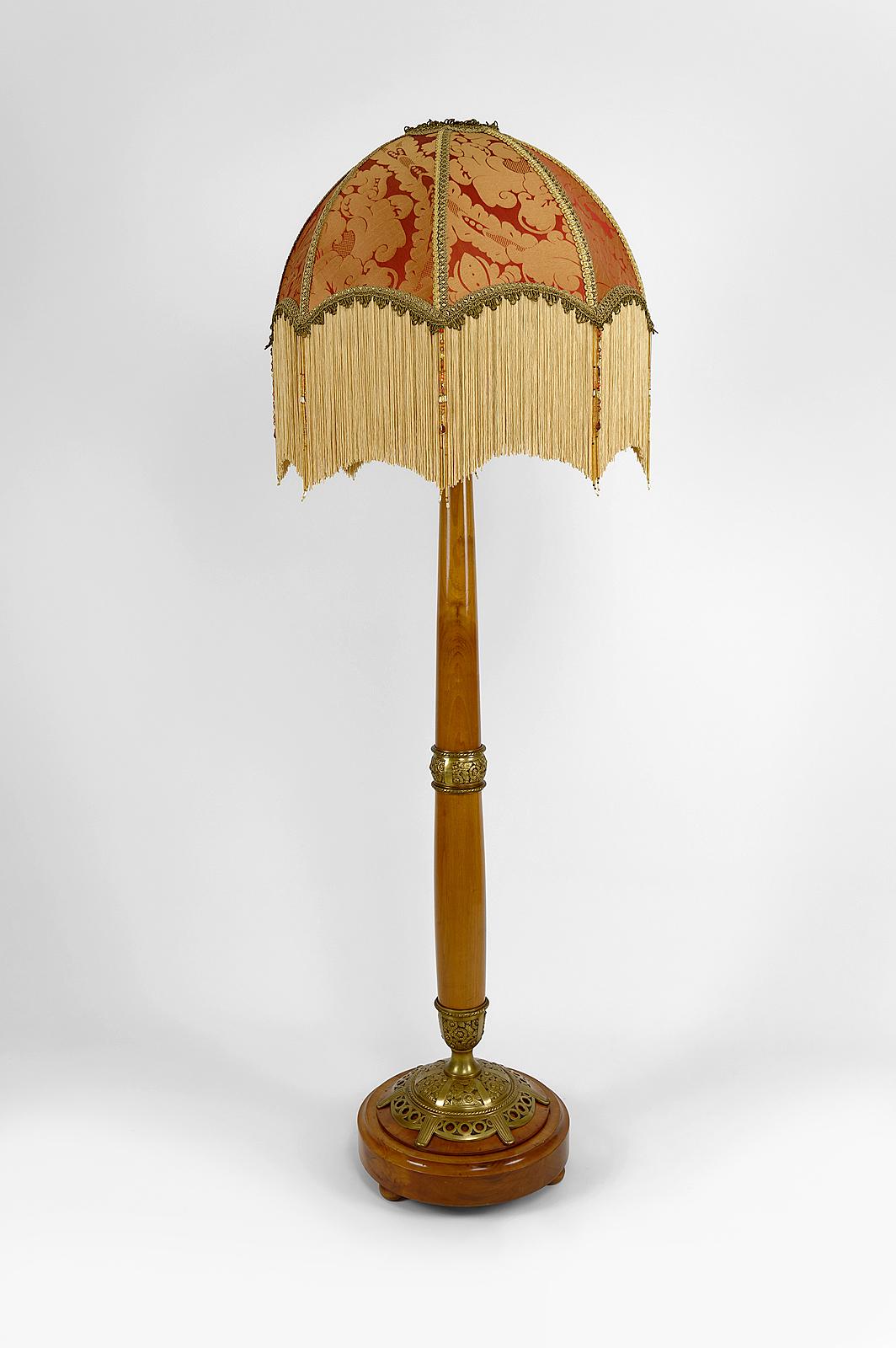 Magnífica lámpara de pie de madera de cerezo rubio compuesta por una base redonda moldeada y un fuste cónico, realzado con elementos decorativos de bronce dorado con motivos florales estilizados.

El conjunto está montado con una pantalla de