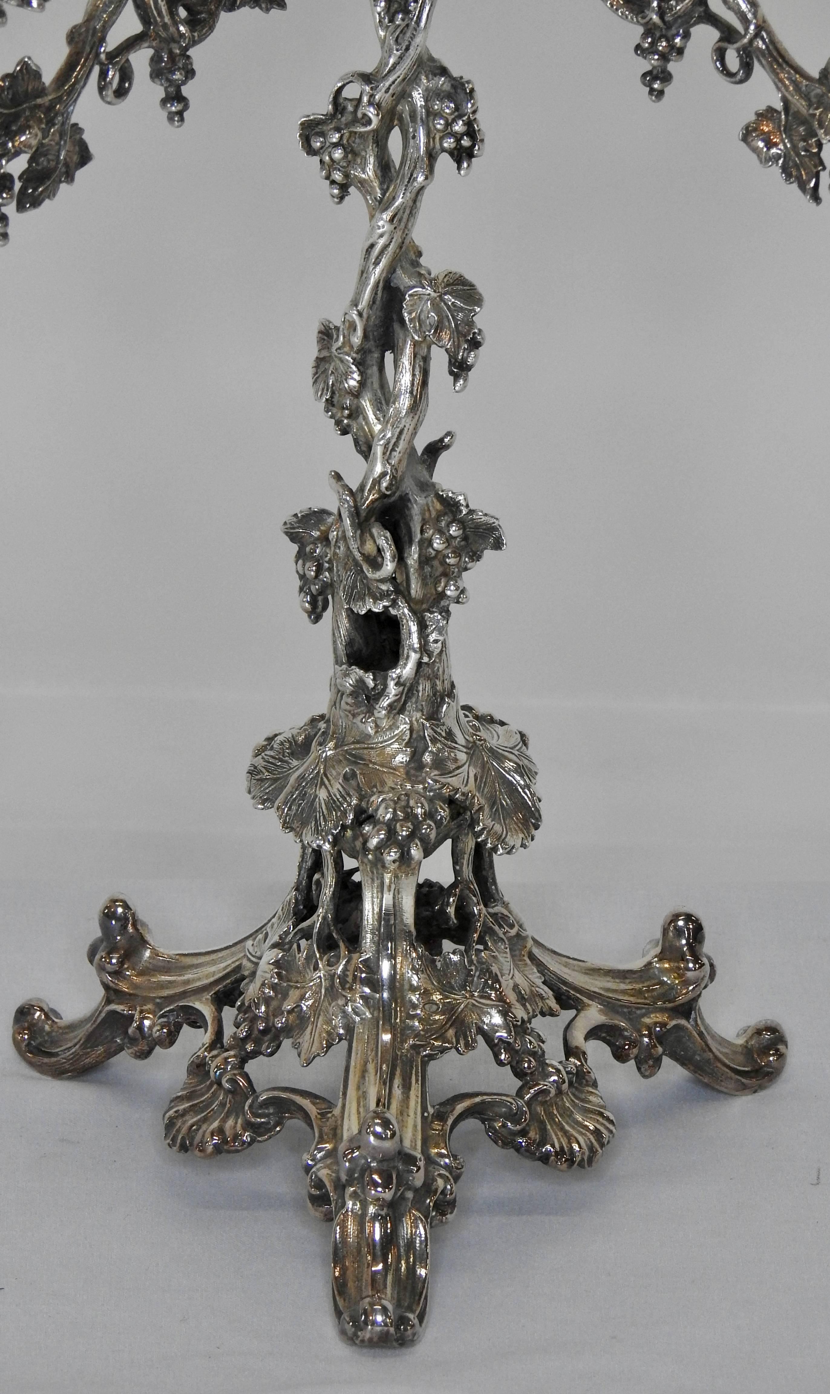 This is a stunning silver plate epergne with four removable arms and an engraved glass bowl. Three ornate feet lead up to vines that twine upward to that arms. The vines twist and go out the arms. From the arms it leads upward to the fluted shape up