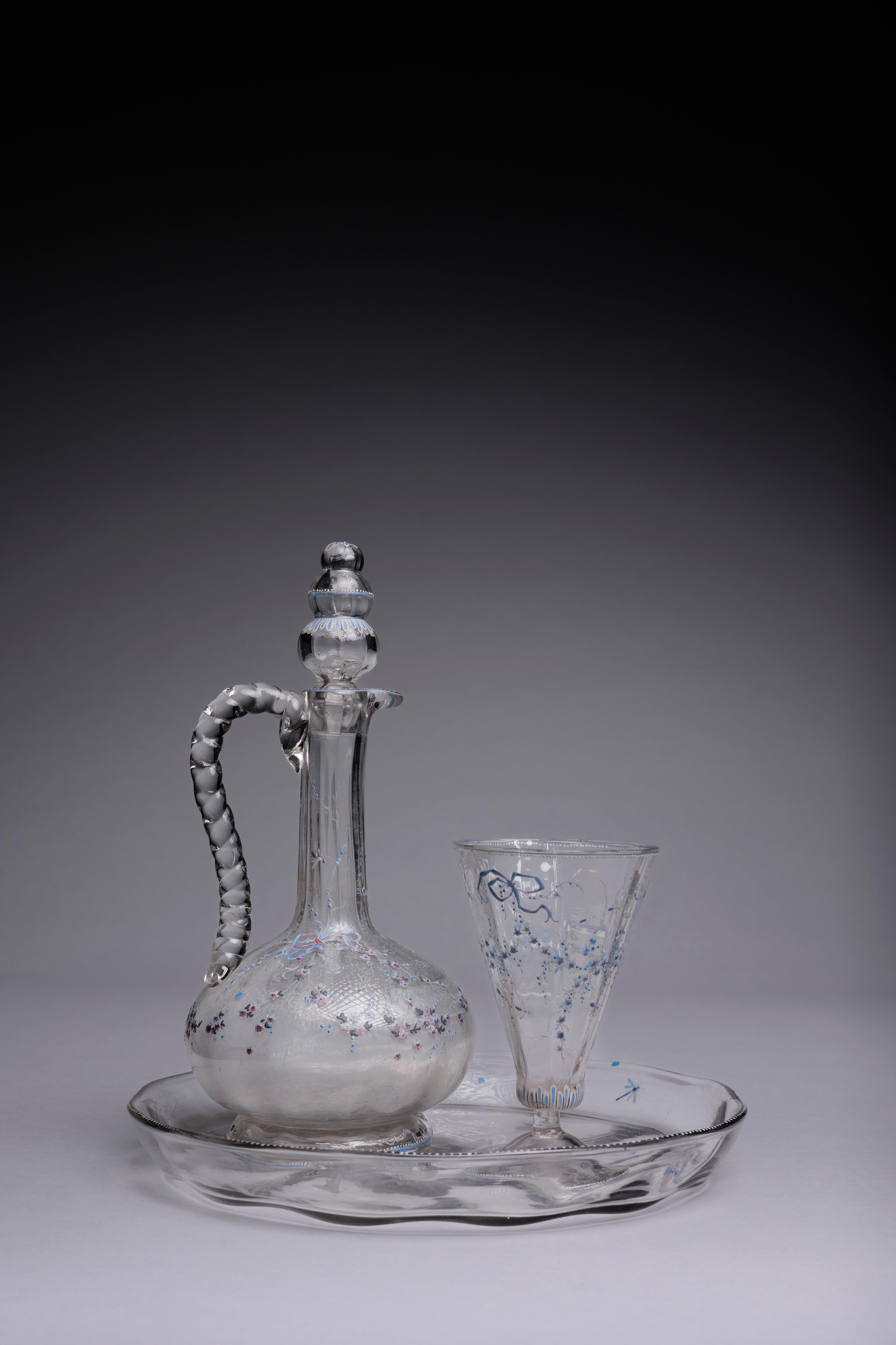 A beautifully delicate French glass decanter set made by Emile Galle in the late 19th century, decorated in enamels with ribbons and flowers in a lovely lavender and periwinkle palette.

This Galle glass set includes a carafe, a footed wine glass,