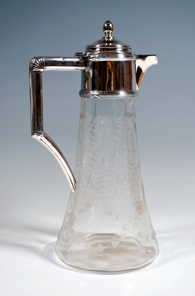 Carafe made of clear glass with a conical body, the inside twelve-point, wave-shaped thickened wall, chiseled floral decoration with feathered leaves and vine leaves, silver fitting with cross ribbon, straight J-shaped handle, covered, beak-shaped