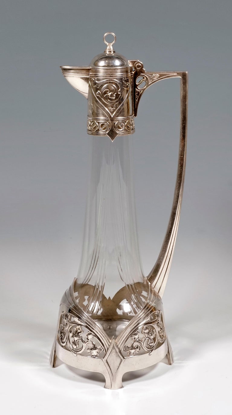 Art Nouveau carafe made of clear glass with a round, upwardly tapering body, front and back two facet bands tapering from bottom to top, the lower, bulbous area enclosed by a fitting of interlocking, profiled bands that enclose openwork leaf
