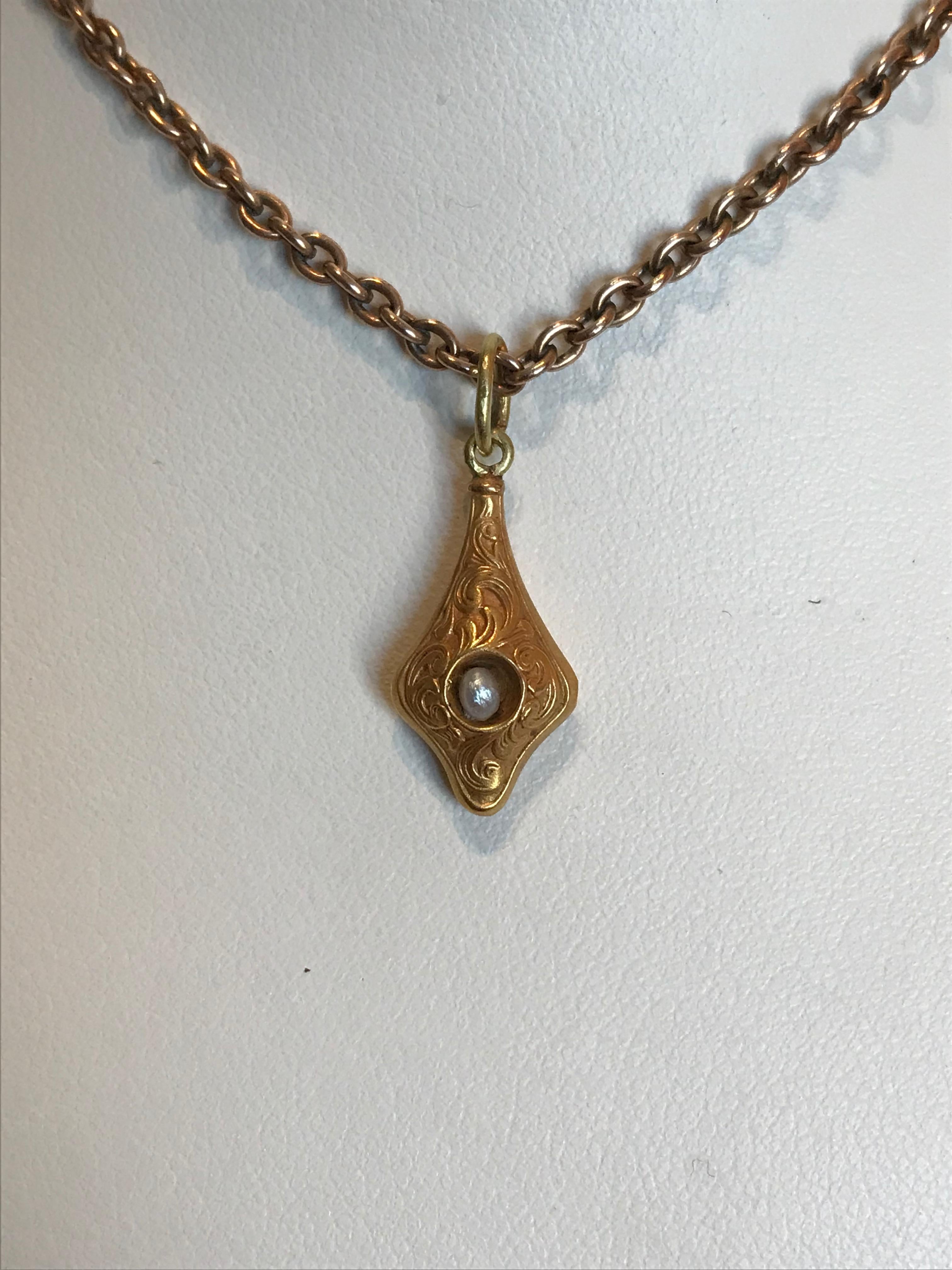 Of 14k matte gold, decorated with raised scrolls and set with a small freshwater natural pearl, with a smooth gold back attached to a gold suspension ring, 

Circa 1900

i in. (2.5 cm) long including suspension ring; 3/8 in. (1 cm) wide; 1/8 in. (.3