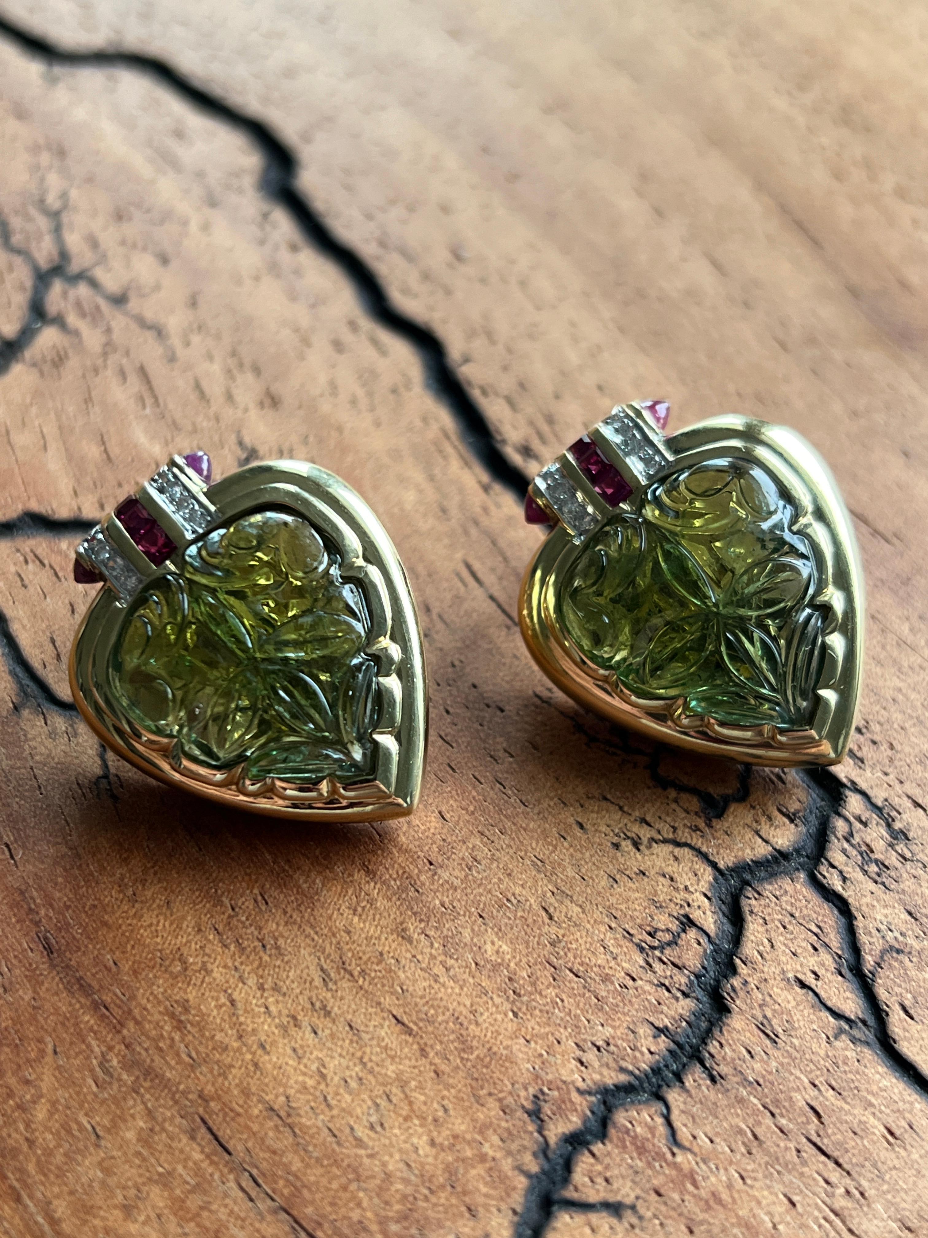 Un paio di orecchini in oro giallo 18 carati con tormaline verdi a forma di vanga o di cuore, intricatamente intagliate al centro e impreziosite da diamanti scintillanti e rubini vibranti. Ogni orecchino è incastonato con sedici diamanti rotondi a